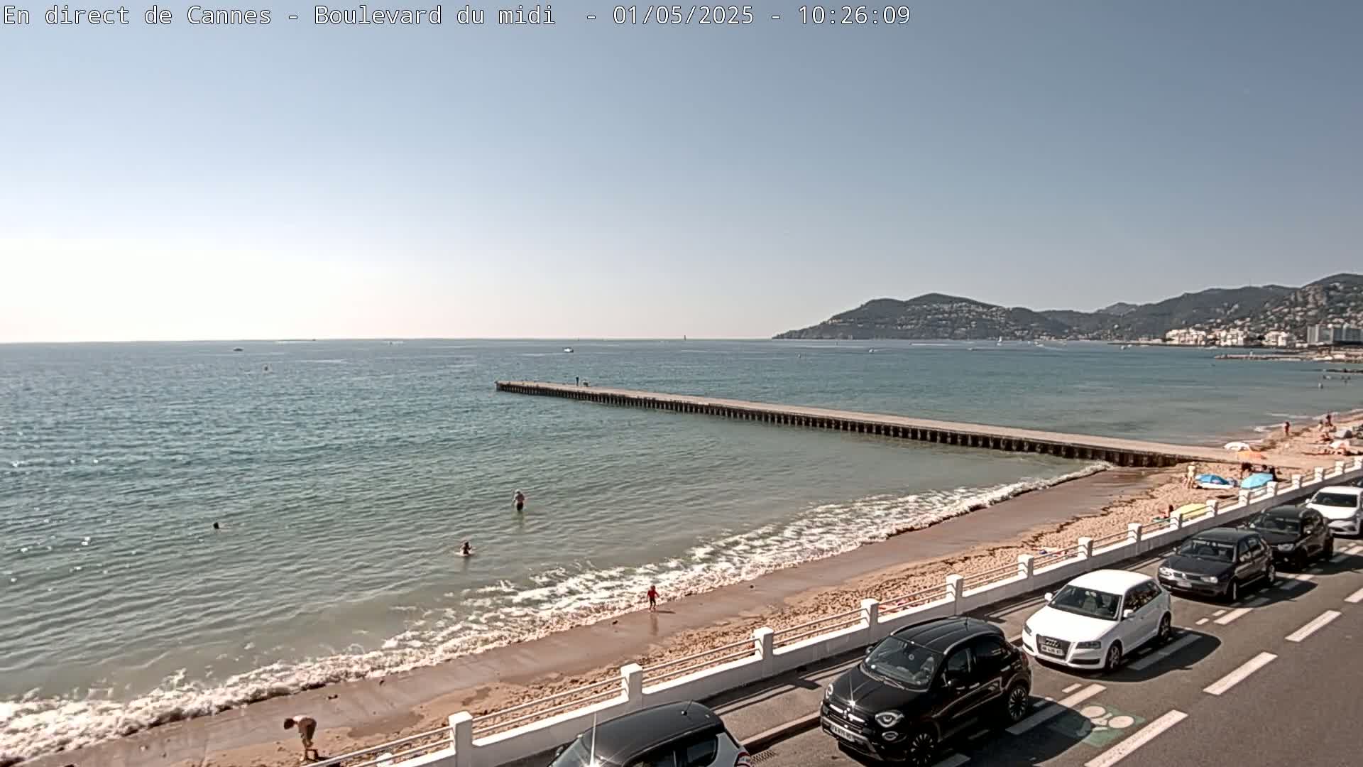 A sandy beach with several people swimming in calm, clear water, a long pier extending into the sea, cars parked along a roadside, and hills in the distance under a sunny sky.
