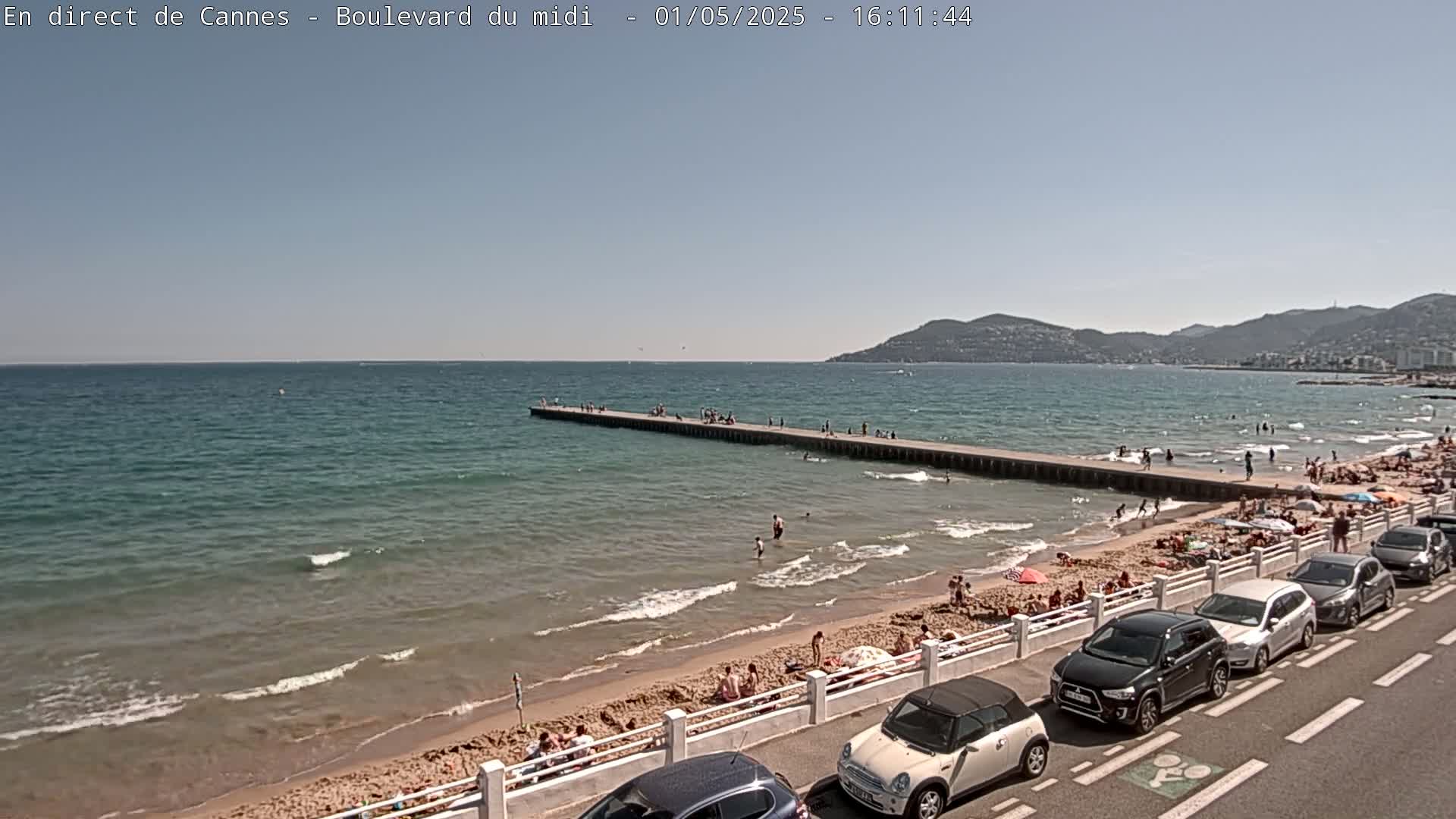 A sunny day at a beach shows a long pier extending into calm, teal water, with people swimming and sunbathing on the shore and parked cars lining a road beside the beach.