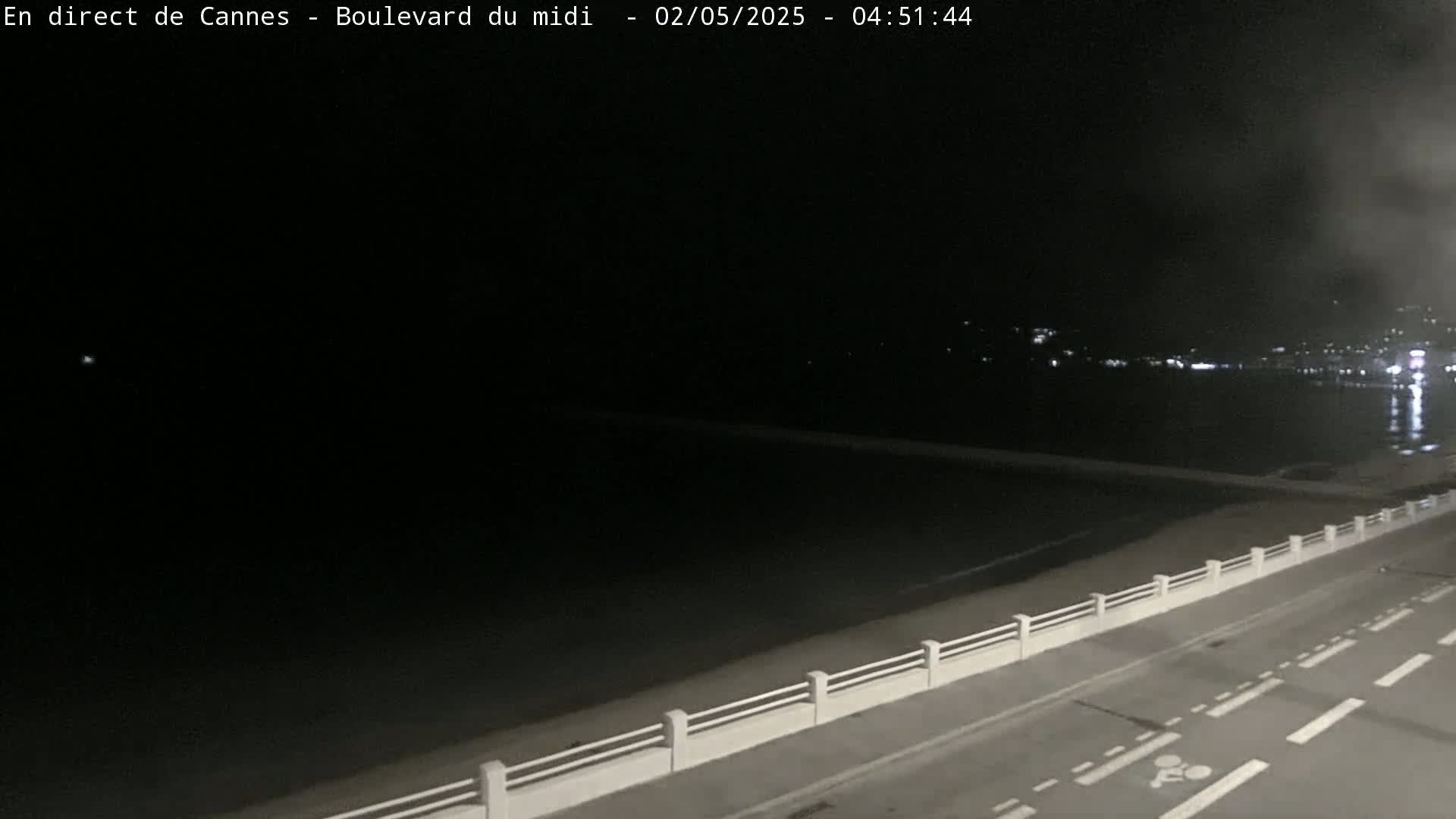 A dark, nighttime view of a road next to a calm sea with a low white railing separating them, and distant lights across the water.