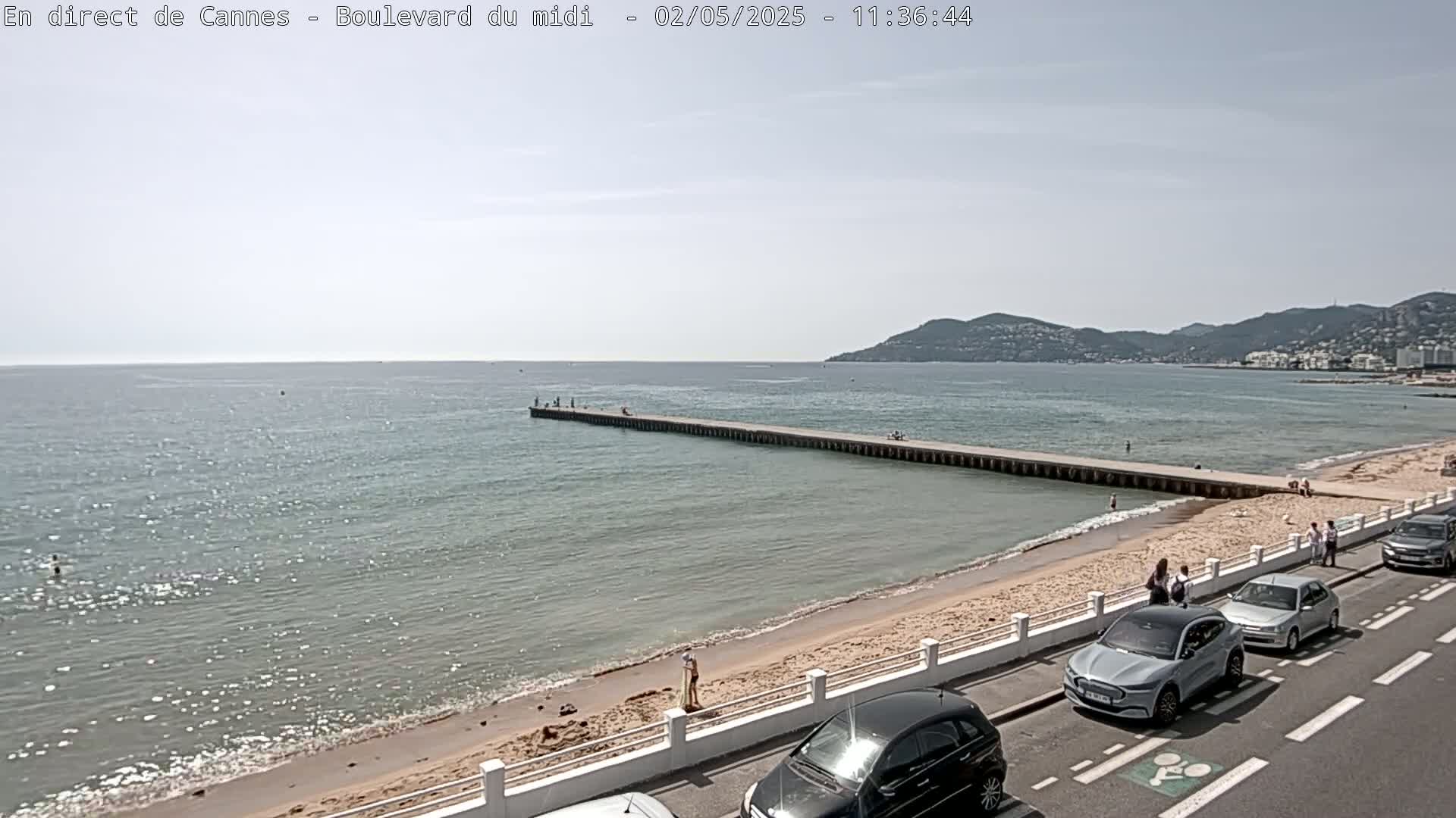 A long pier extends into a calm, light-blue sea under a mostly sunny sky, with a sandy beach and parked cars lining a road beside it.