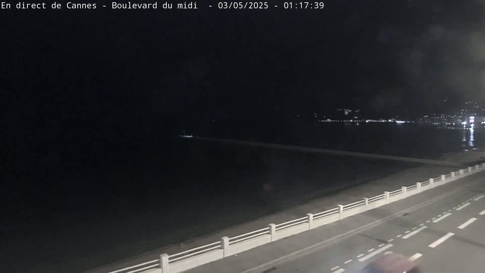 A nighttime, long shot of a dark road next to a beach and calm water with city lights in the distance.