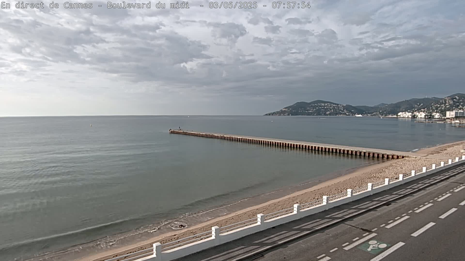 A long pier extends into a calm sea under a cloudy sky, beside a sandy beach and a paved road.