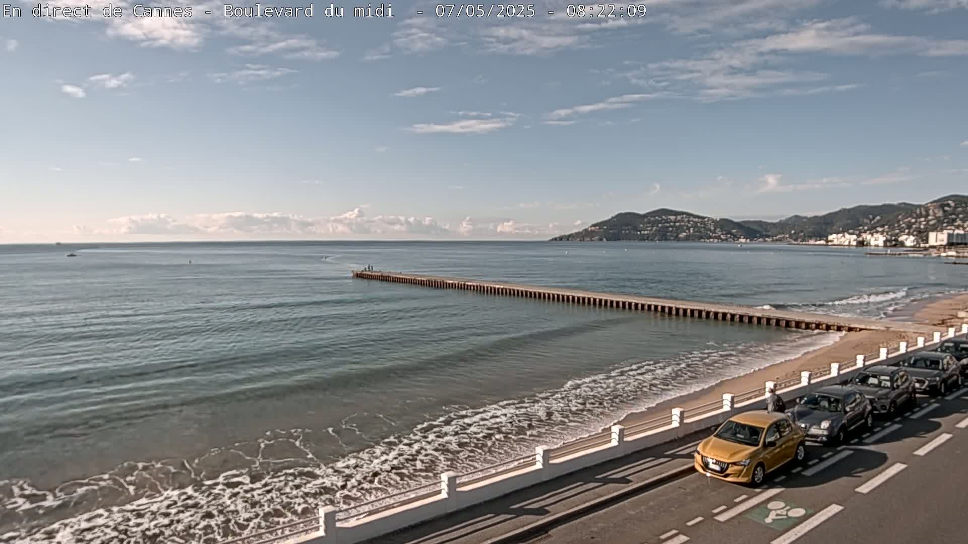 A long pier extends into a calm sea with small waves lapping a sandy beach, where several cars are parked along a road next to a low white wall.
