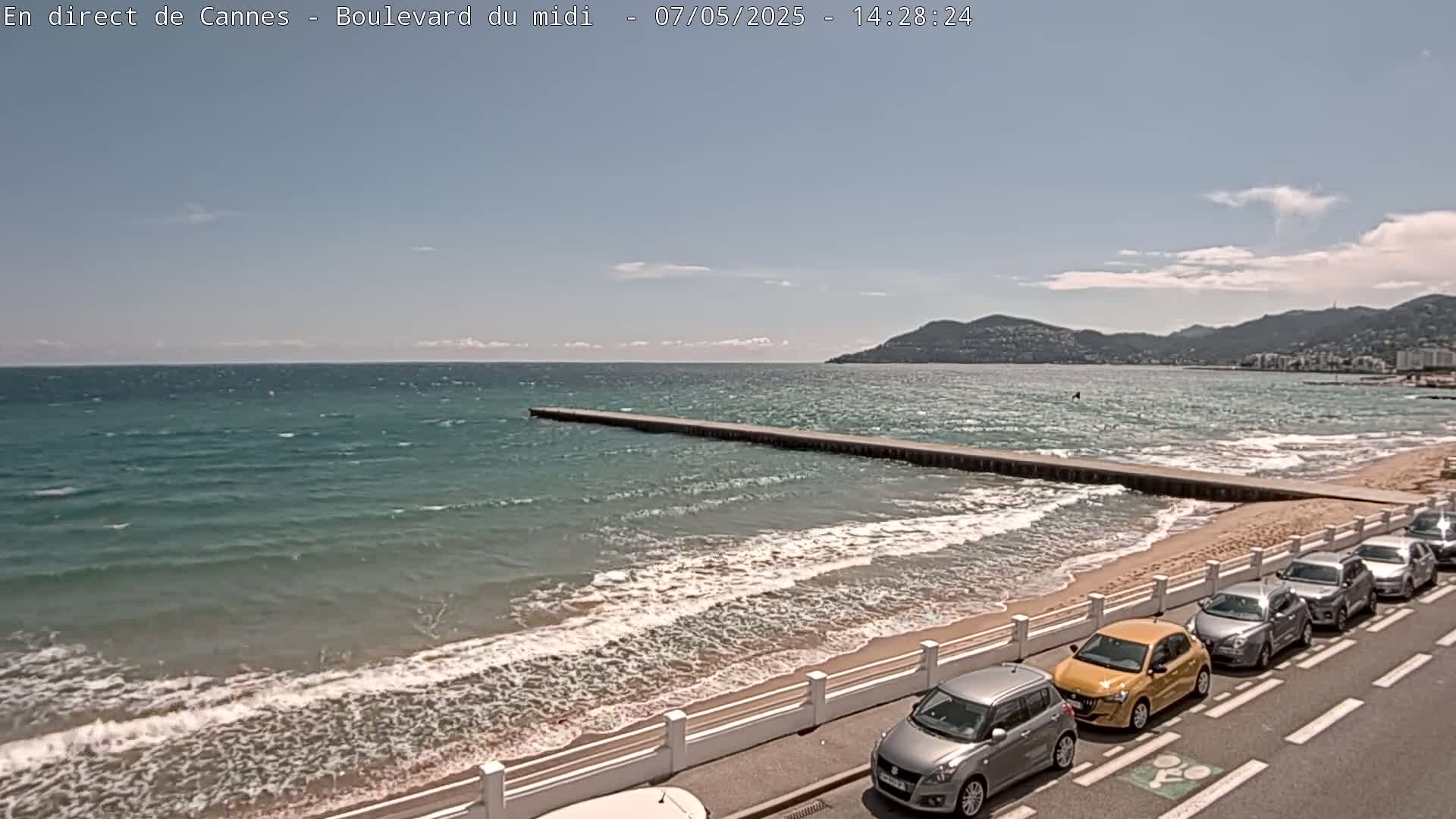 A sandy beach with a long pier extends into a wavy turquoise sea under a partly cloudy sky, with several cars parked along the roadside.