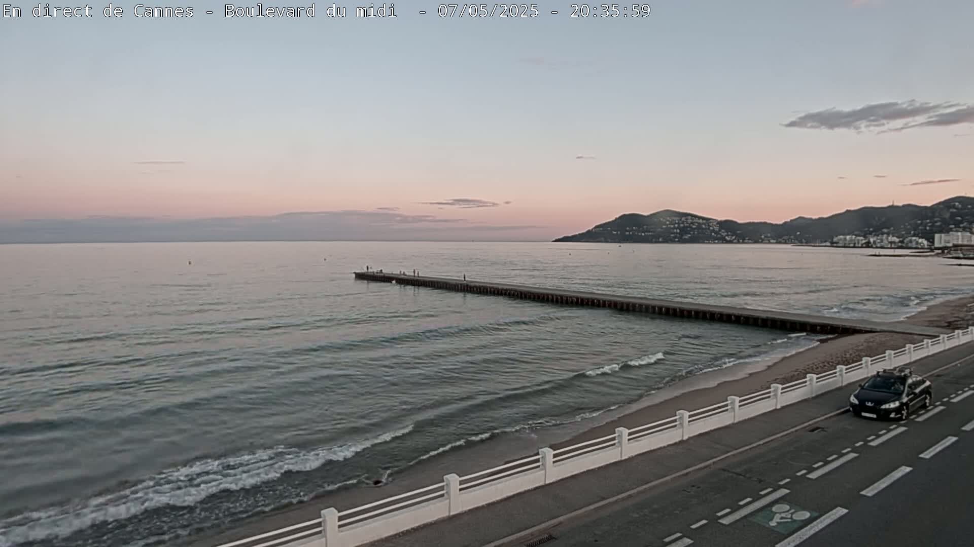 A long pier extends into a calm sea at sunset, with a car parked on a nearby road beside a white fence.