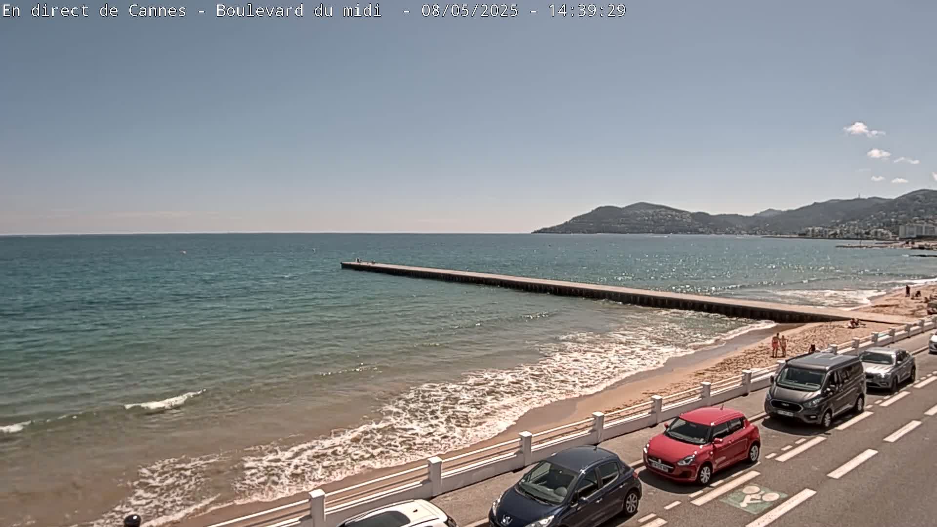 A long pier extends from a sandy beach into a calm, blue ocean under a clear, sunny sky, with cars parked along the road bordering the beach.