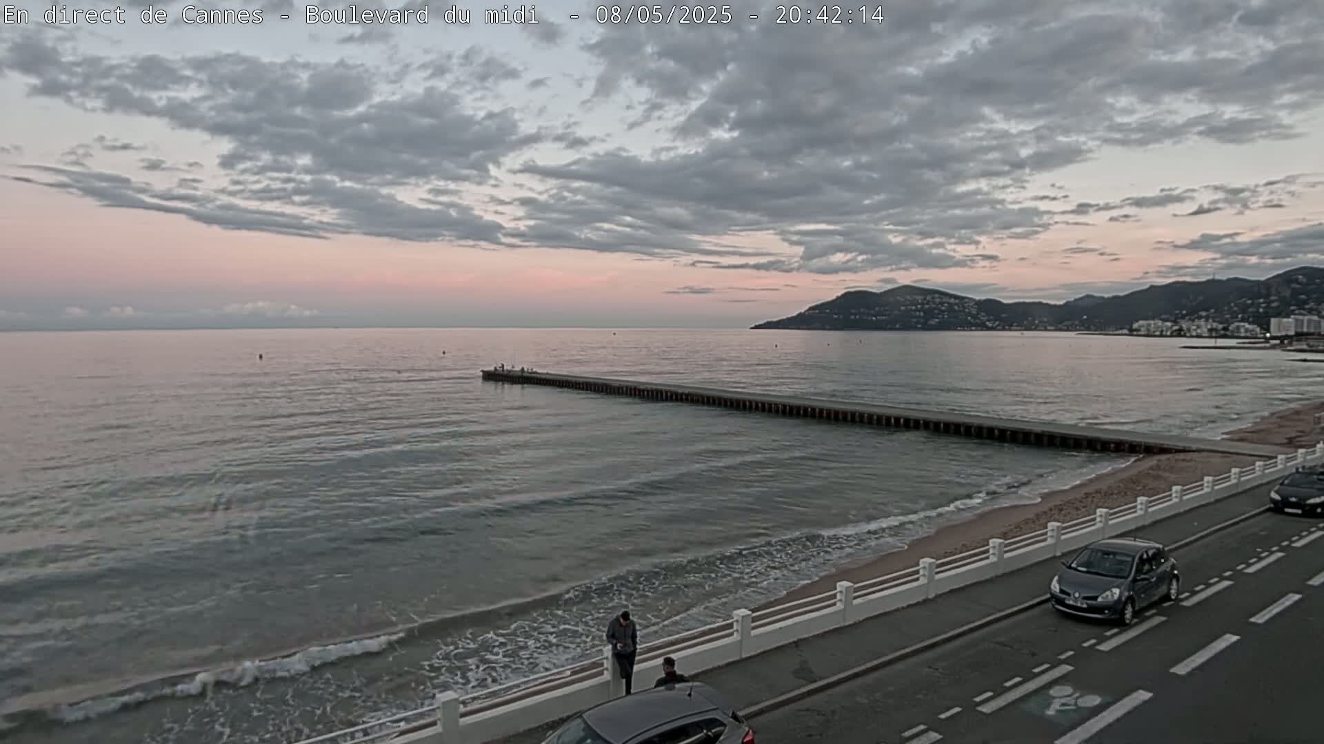 A long pier extends into a calm sea under a partly cloudy, pinkish sunset sky, with a beach and road visible in the foreground.