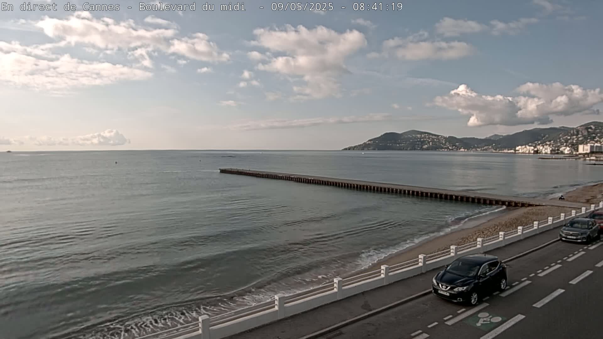 A partly cloudy day shows a long pier extending into a calm sea, with a sandy beach and road alongside, and several cars parked along the roadside.
