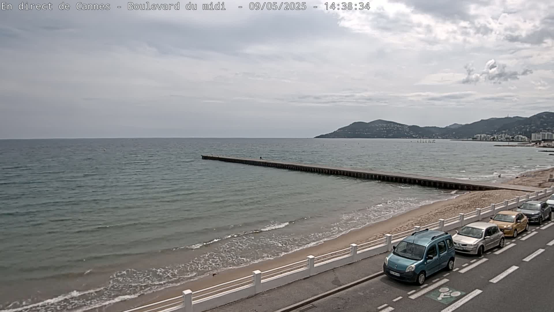 A sandy beach with calm, light-grey water, a long pier extending into the sea, several cars parked along a roadside, and distant mountains under a cloudy sky.