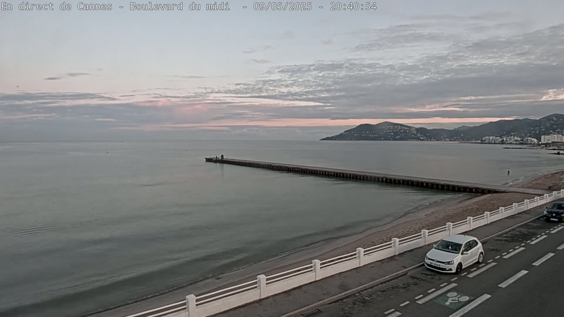 A long pier extends into a calm sea under a cloudy, pinkish-hued sky, with a beach and a road visible in the foreground.