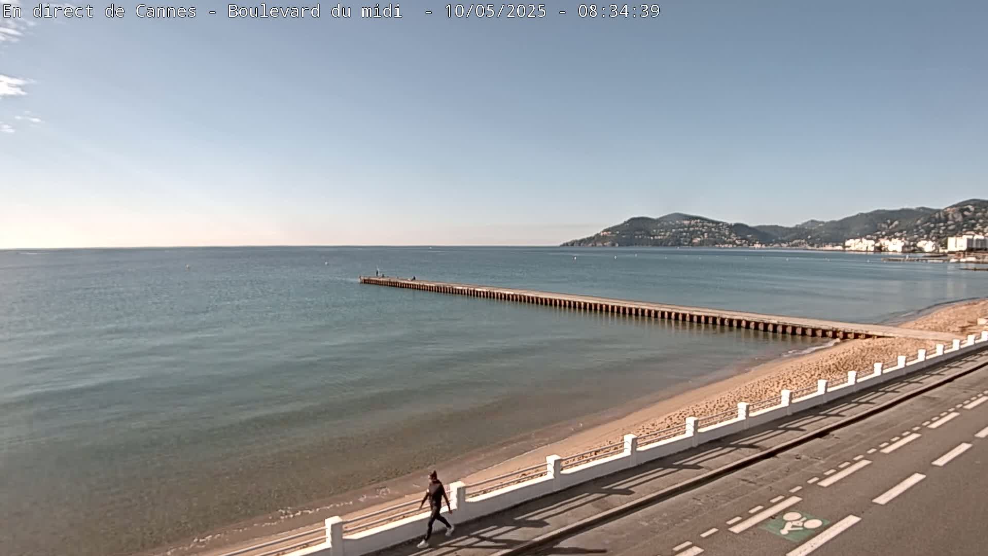 A person walks along a paved road next to a sandy beach and calm ocean with a long pier extending into the water under a clear sky.