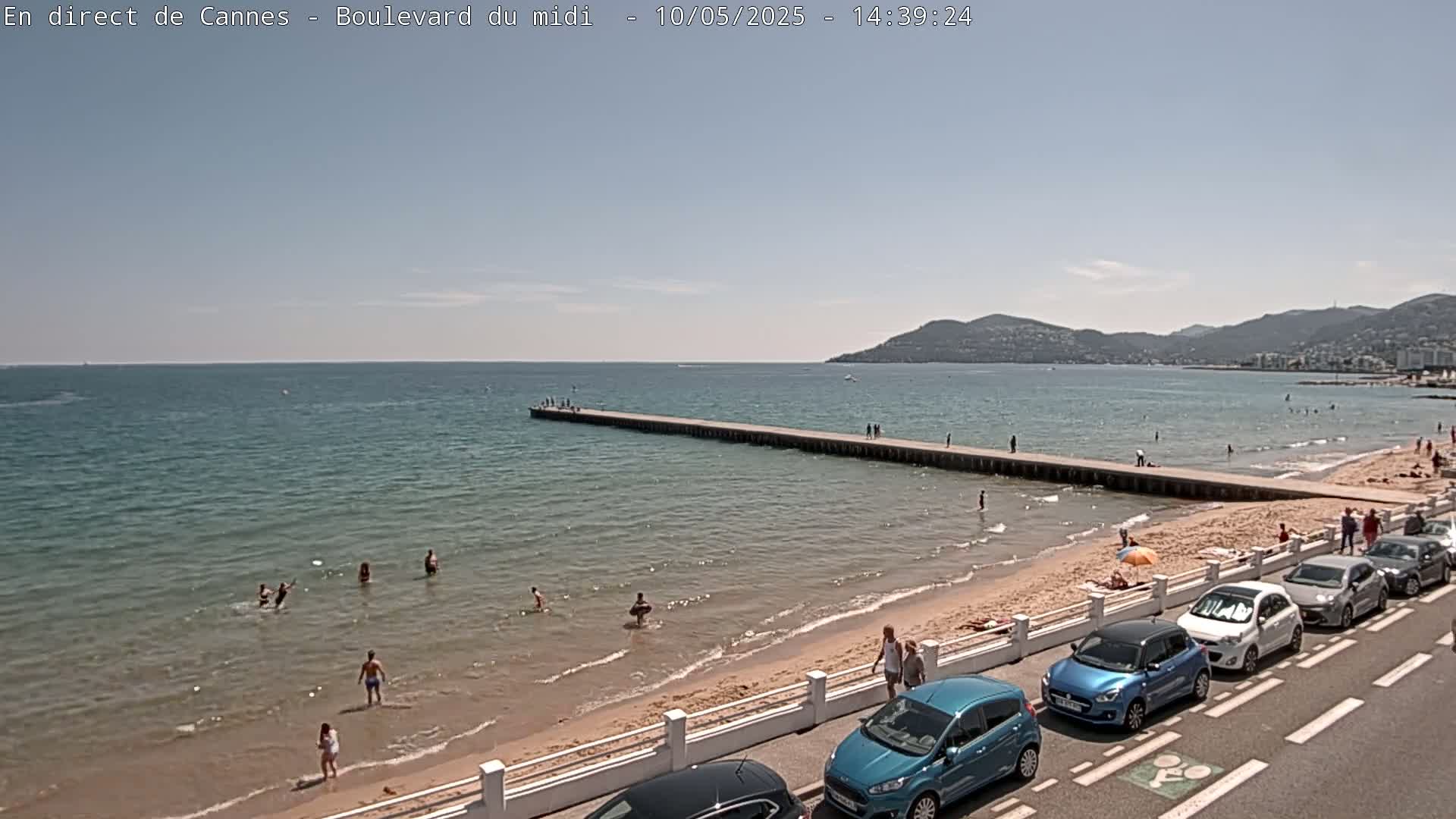 A sandy beach with people swimming and sunbathing, a long pier extending into a calm blue sea, and cars parked along a roadside under a clear sunny sky.