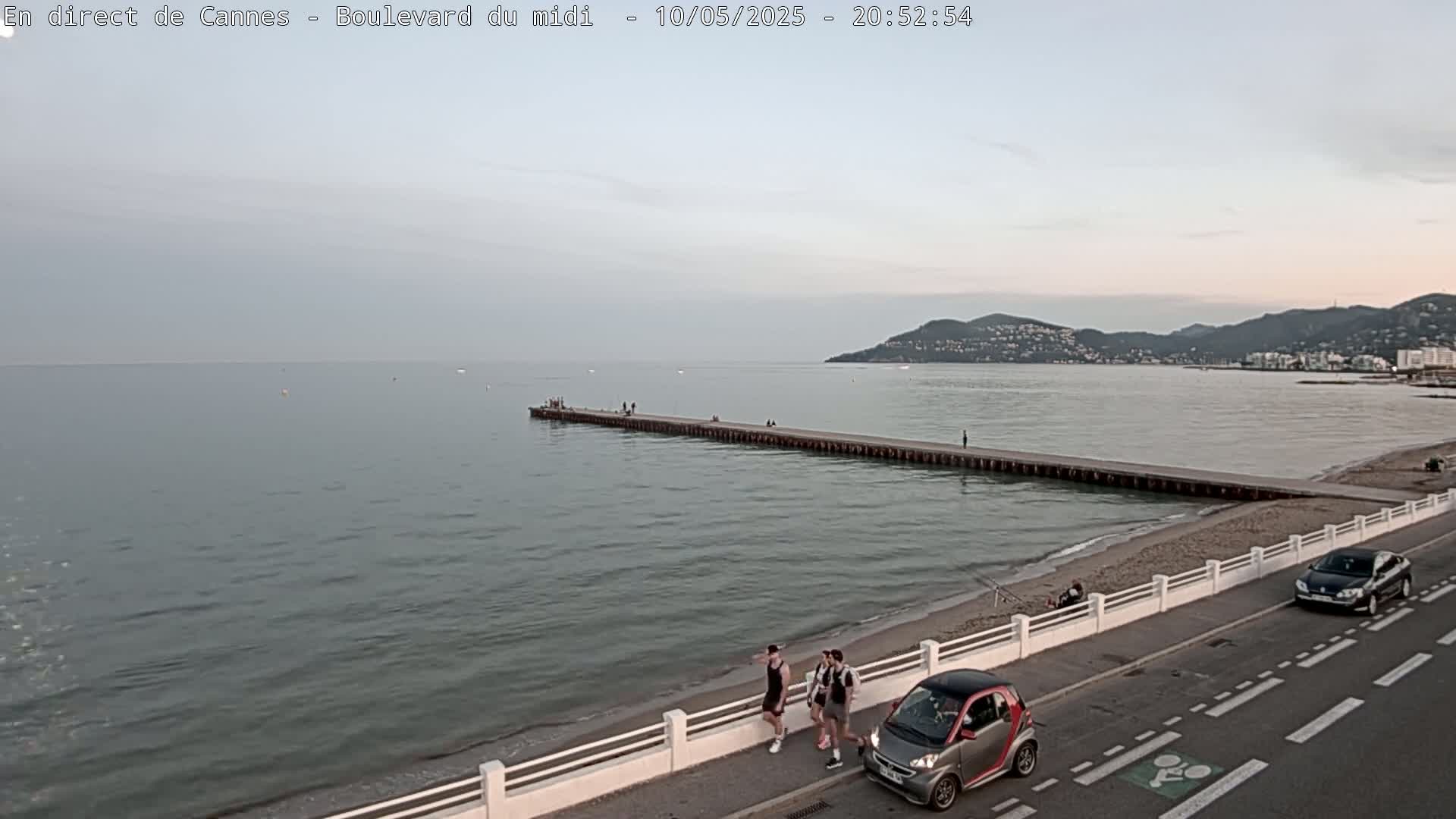 A long pier extends into a calm sea under a twilight sky, with a few people walking along a beach and a road beside it, and cars parked nearby.