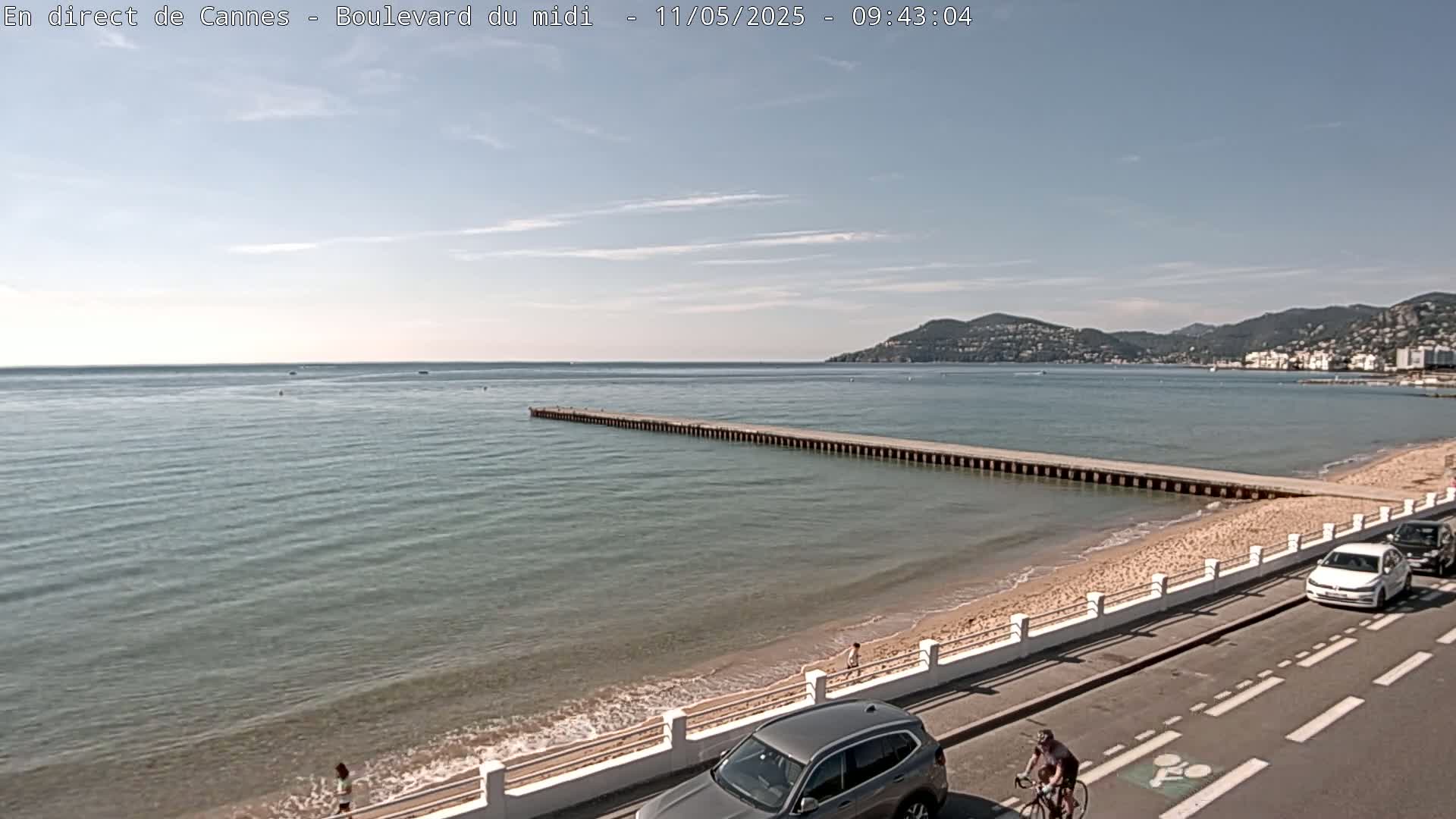 A long pier extends into a calm, light-blue sea next to a sandy beach, with cars parked along a road overlooking the water under a mostly clear sky.