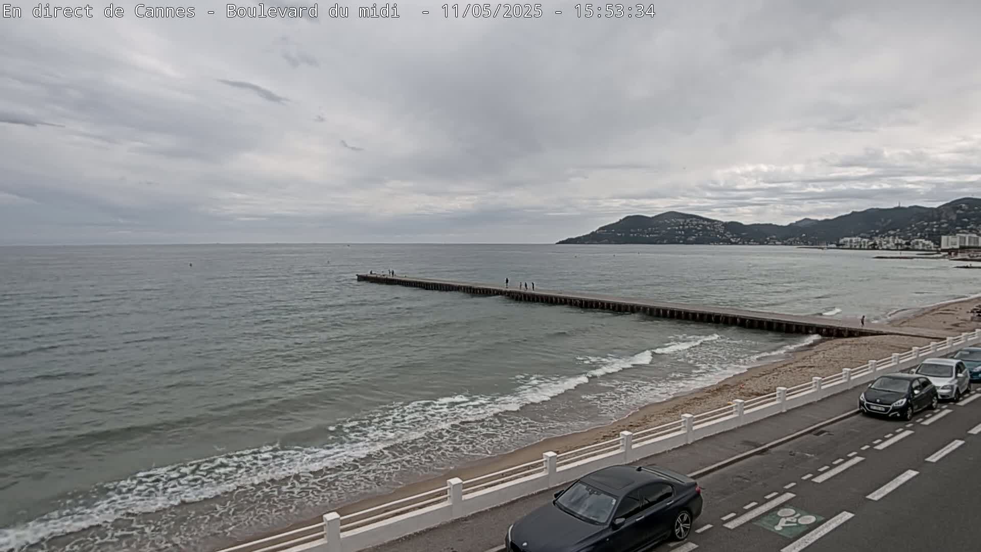 A long pier extends into a calm, gray sea under an overcast sky, with a sandy beach and a road lined with parked cars visible in the foreground.