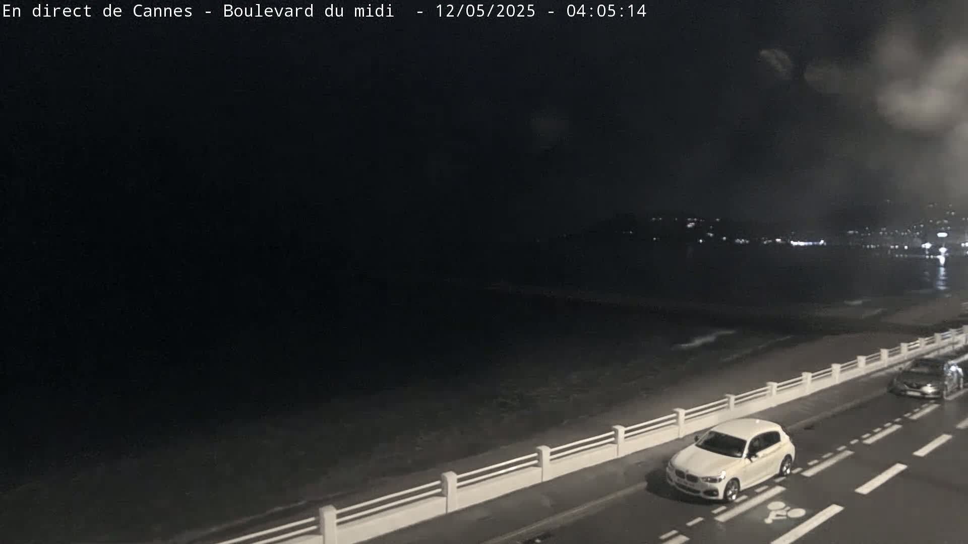 A nighttime, seaside view shows a road next to a low wall overlooking a dark ocean, with a white car parked on the road and another further down.