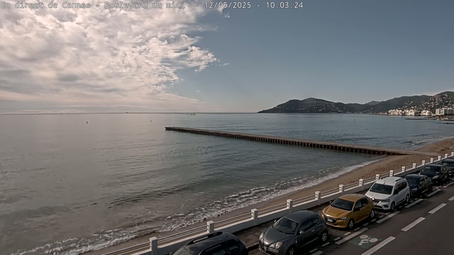 A partly cloudy day shows a calm sea with a long jetty extending into the water, a sandy beach lined with parked cars, and distant hills.