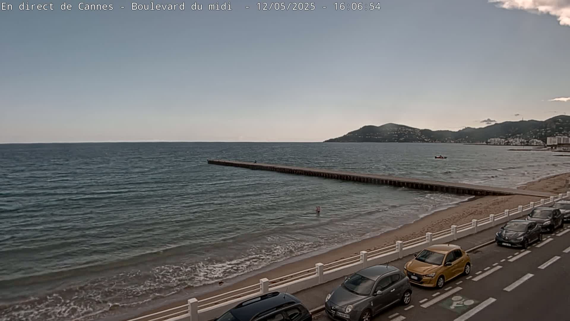 A sandy beach with parked cars along a roadway next to a calm sea and a long pier extending into the water under a mostly cloudy sky.
