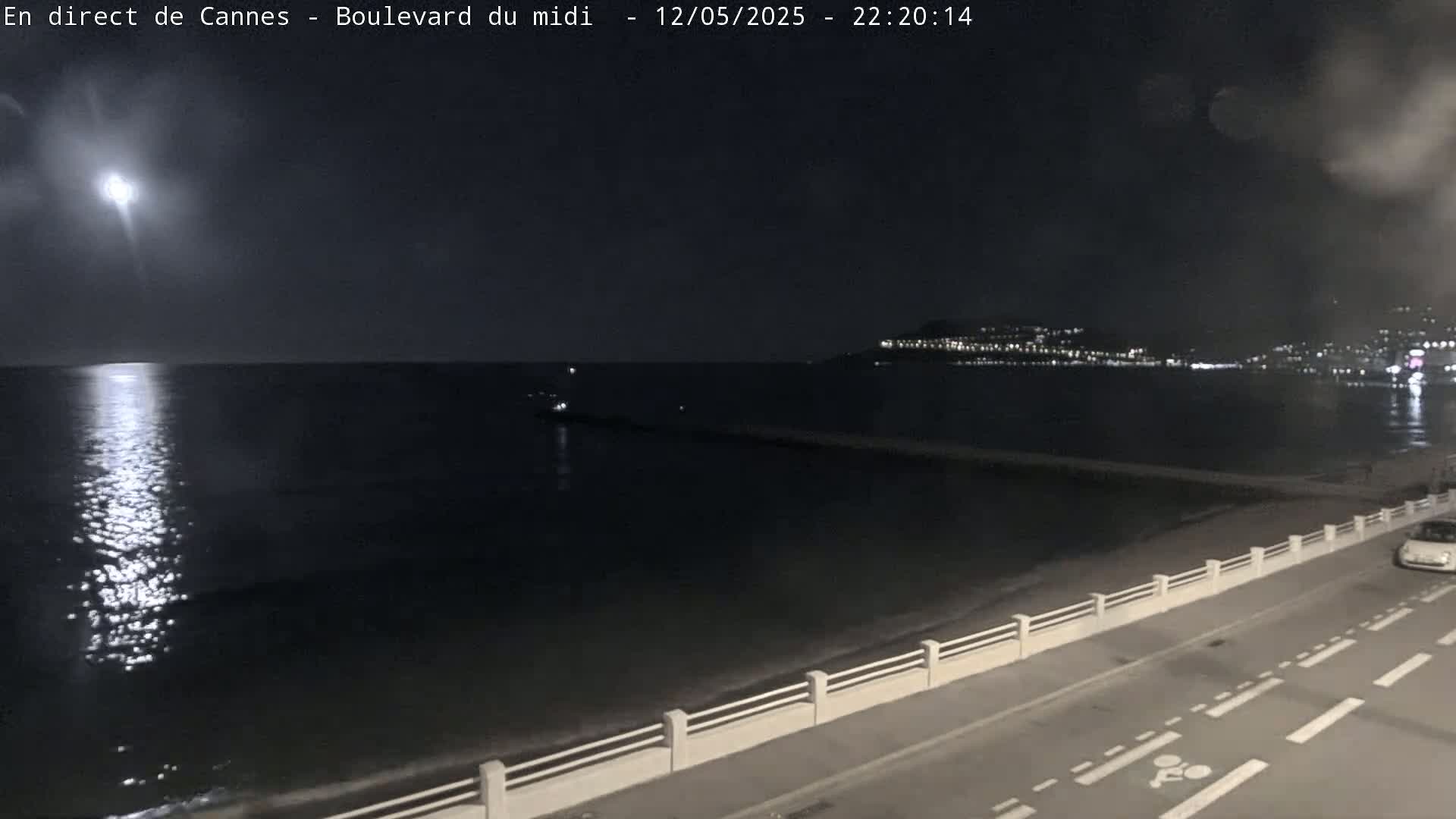 A bright moon shines over a dark, calm sea at night, illuminating a shoreline road with a low white railing and a distant city with lights.