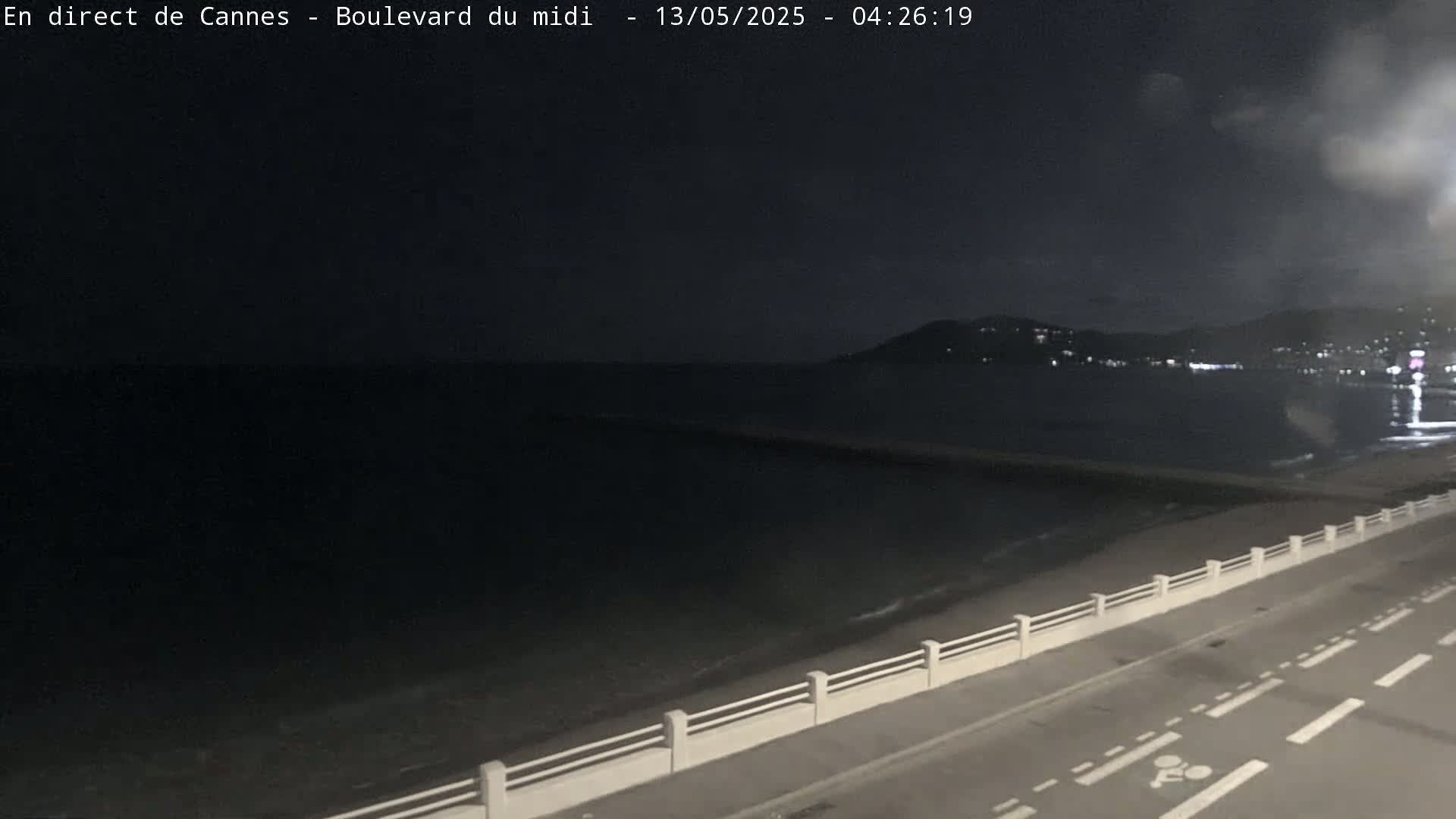 A dark, nighttime view of a calm sea with a road and low wall along the shore, and distant lights visible on the opposite coast.
