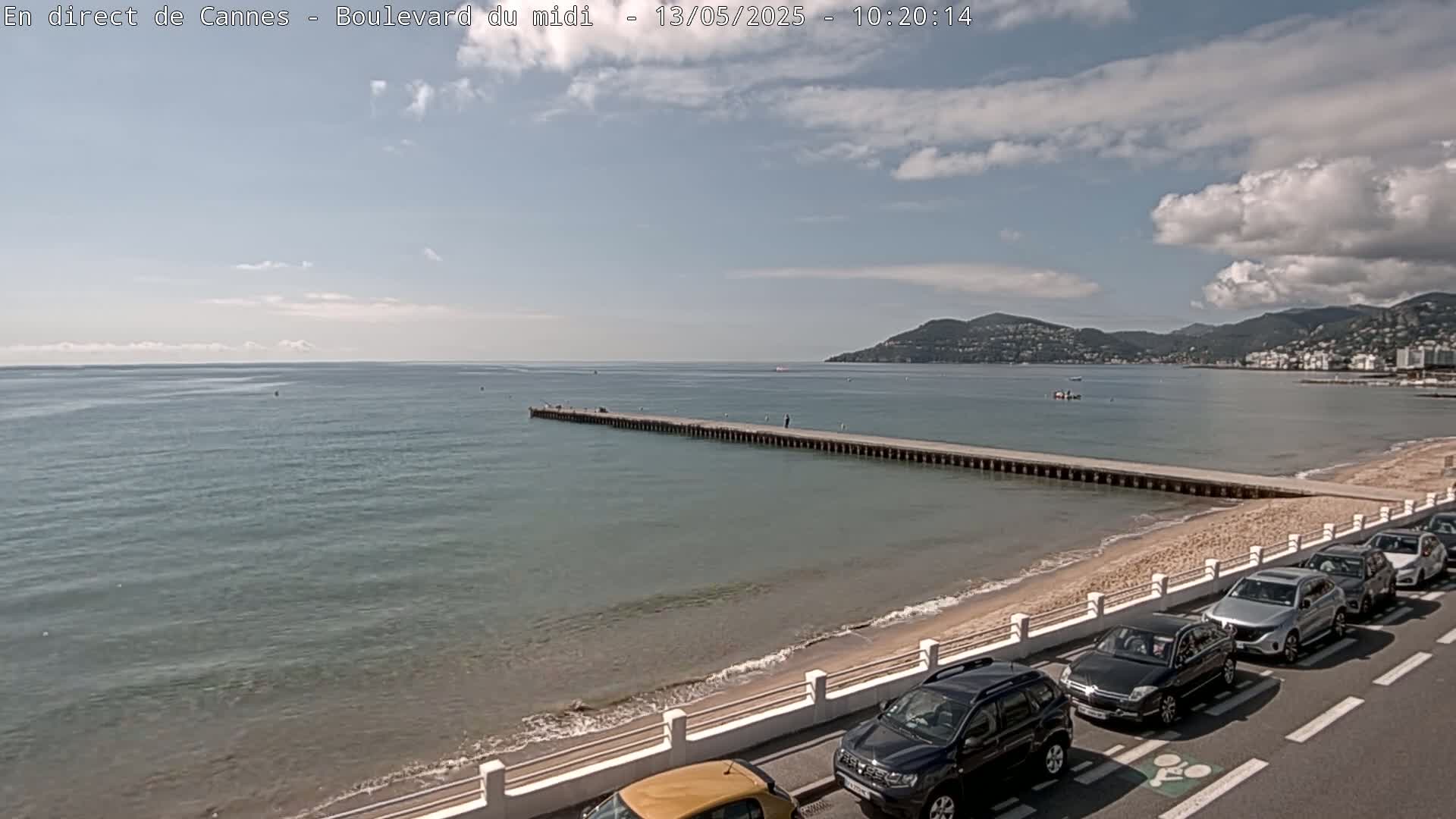 A long pier extends into a calm, blue sea under a partly cloudy sky, with a sandy beach and parked cars lining a road beside it.