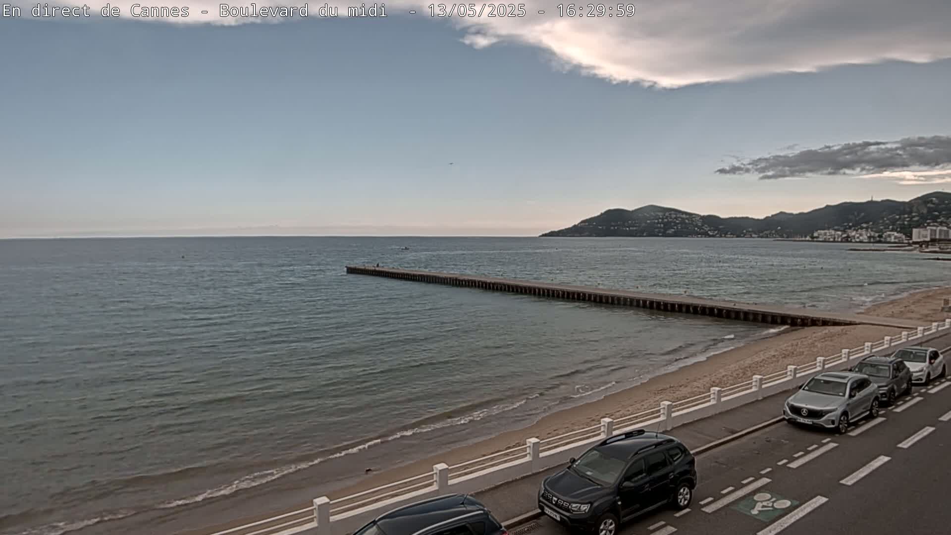 A long pier extends into a calm sea under a partly cloudy sky, with several cars parked along a road next to a sandy beach and distant hills.