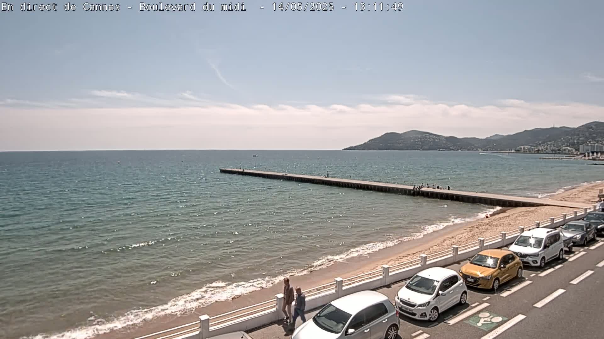 A long pier extends into a calm sea next to a sandy beach where several cars are parked along a roadside, under a mostly sunny sky.