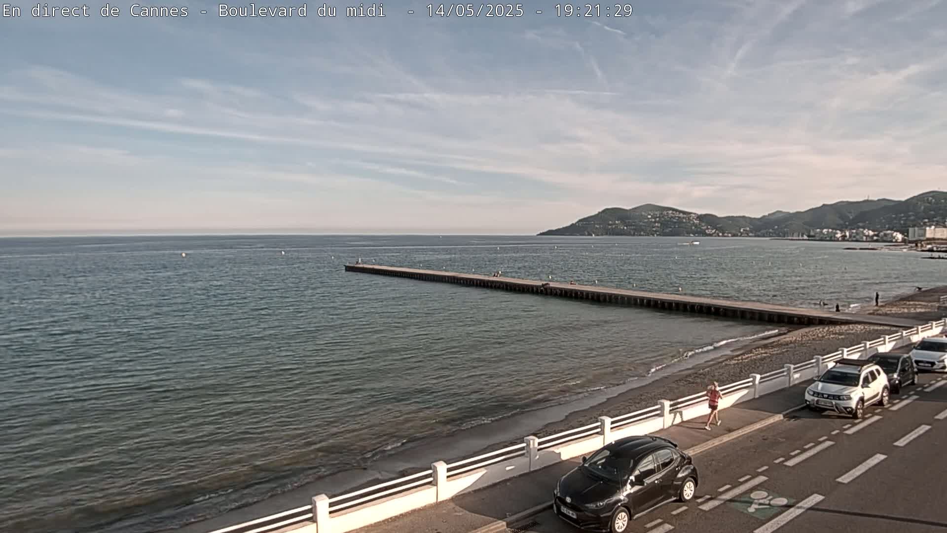 A long pier extends into a calm sea under a partly cloudy sky, with a beach, road, and parked cars visible along the shore.