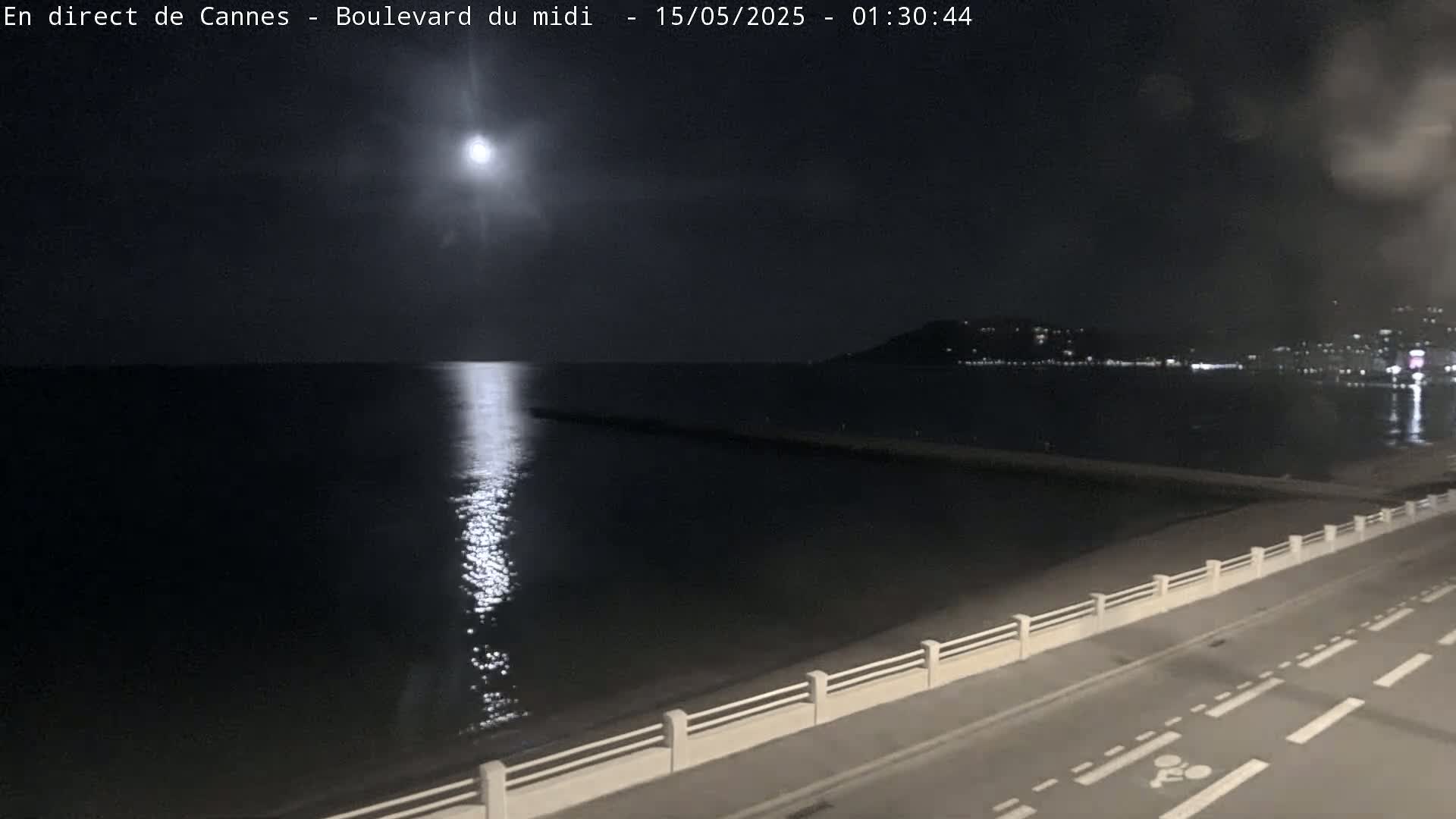 A bright moon illuminates a dark, calm sea at night, reflecting on the water near a paved road and low wall.