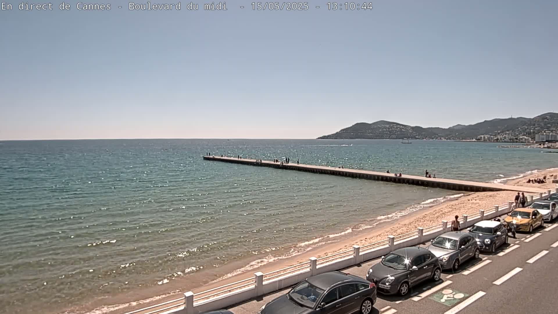 A long pier extends from a sandy beach into a calm, clear ocean under a sunny sky, with several cars parked along the roadside.