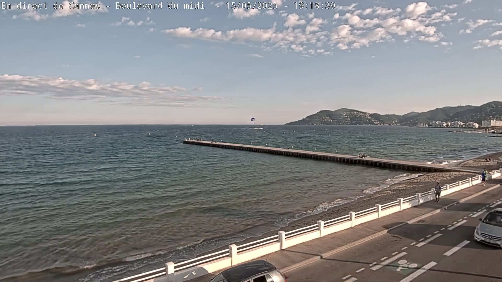 A long pier extends into a calm, blue sea under a partly cloudy sky, with a sandy beach and a road visible along the shore, and mountains in the distance.