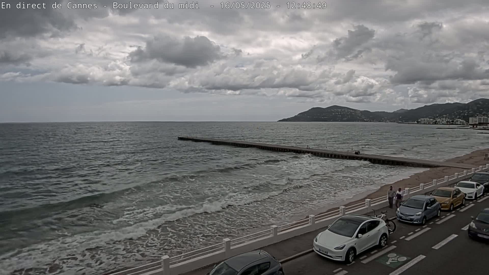 A cloudy day at a beach with moderate waves, a long pier, and cars parked along a roadside.