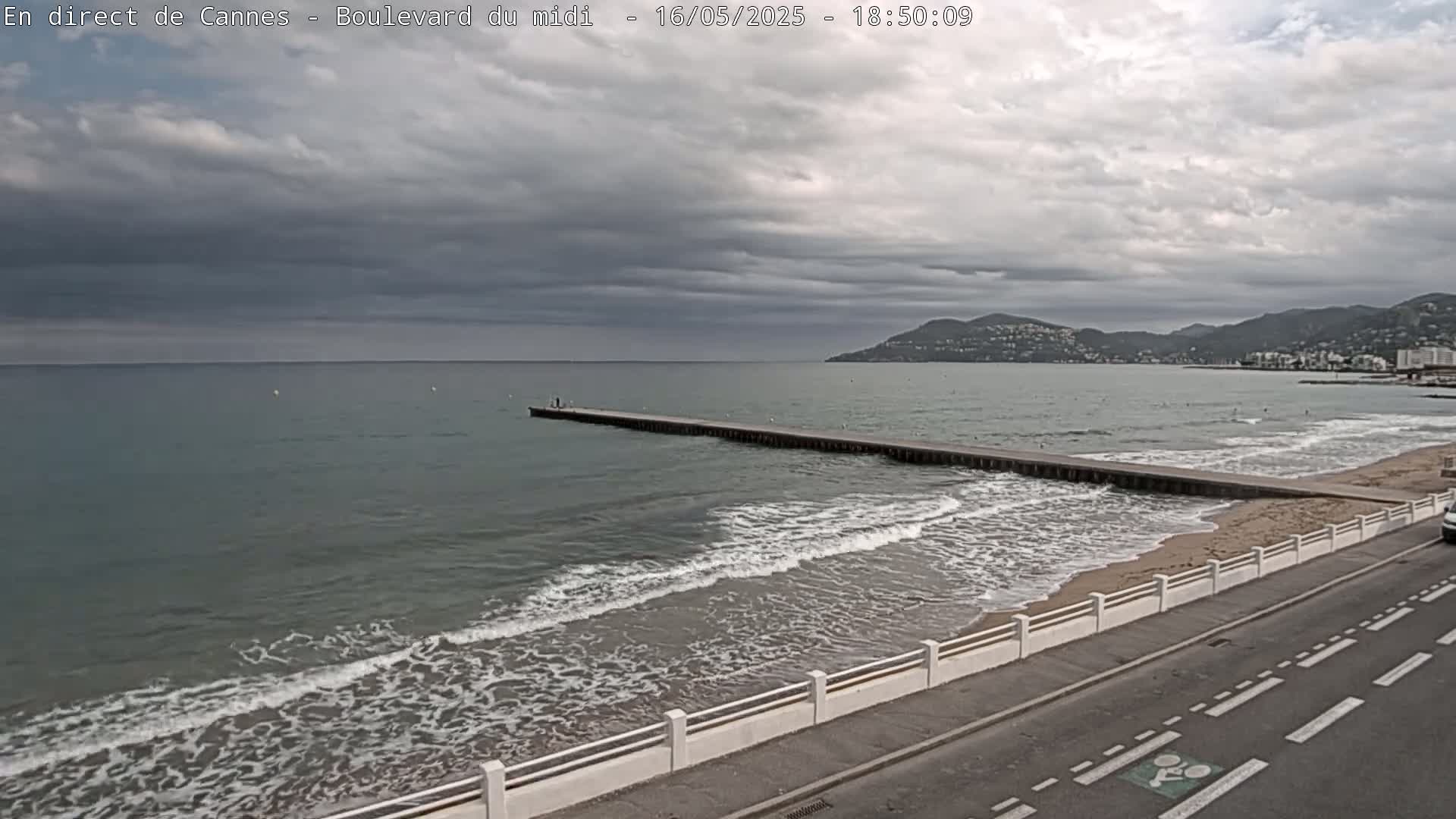 A long pier extends into a calm sea with small waves lapping a sandy beach under an overcast sky.