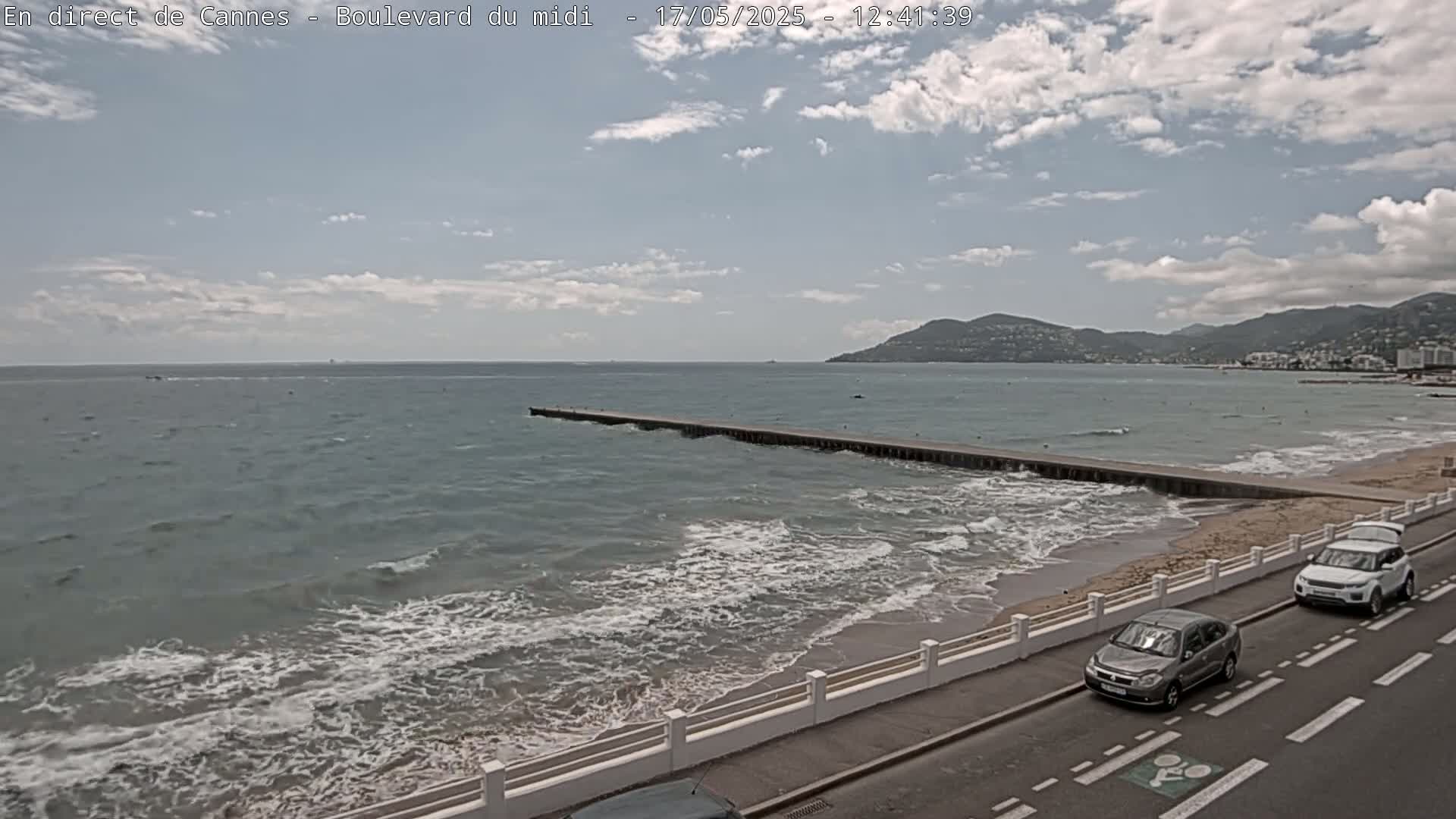 A partly cloudy day shows a wavy ocean with a long pier extending into it, along a sandy beach with a road and cars nearby.