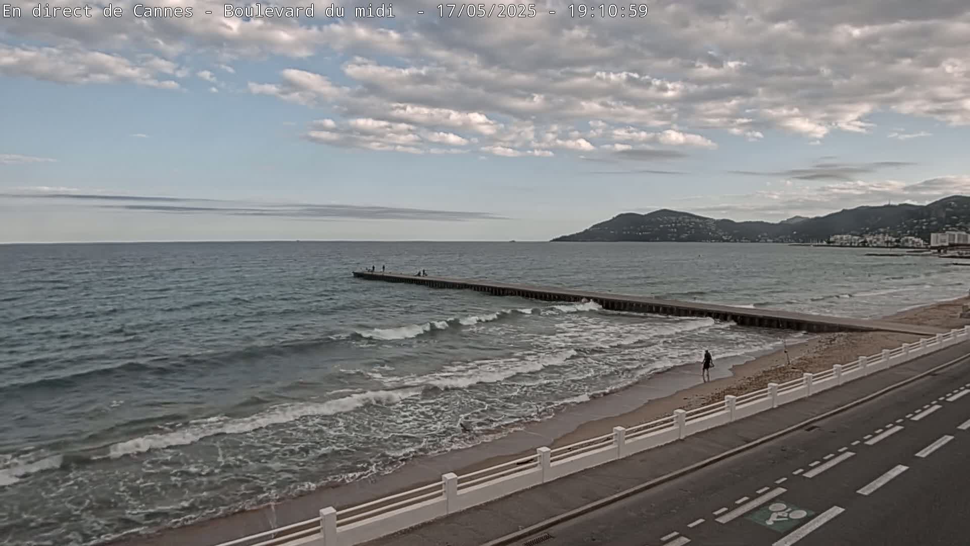 A long pier extends into a wavy sea under a partly cloudy sky, with a sandy beach and road visible in the foreground.