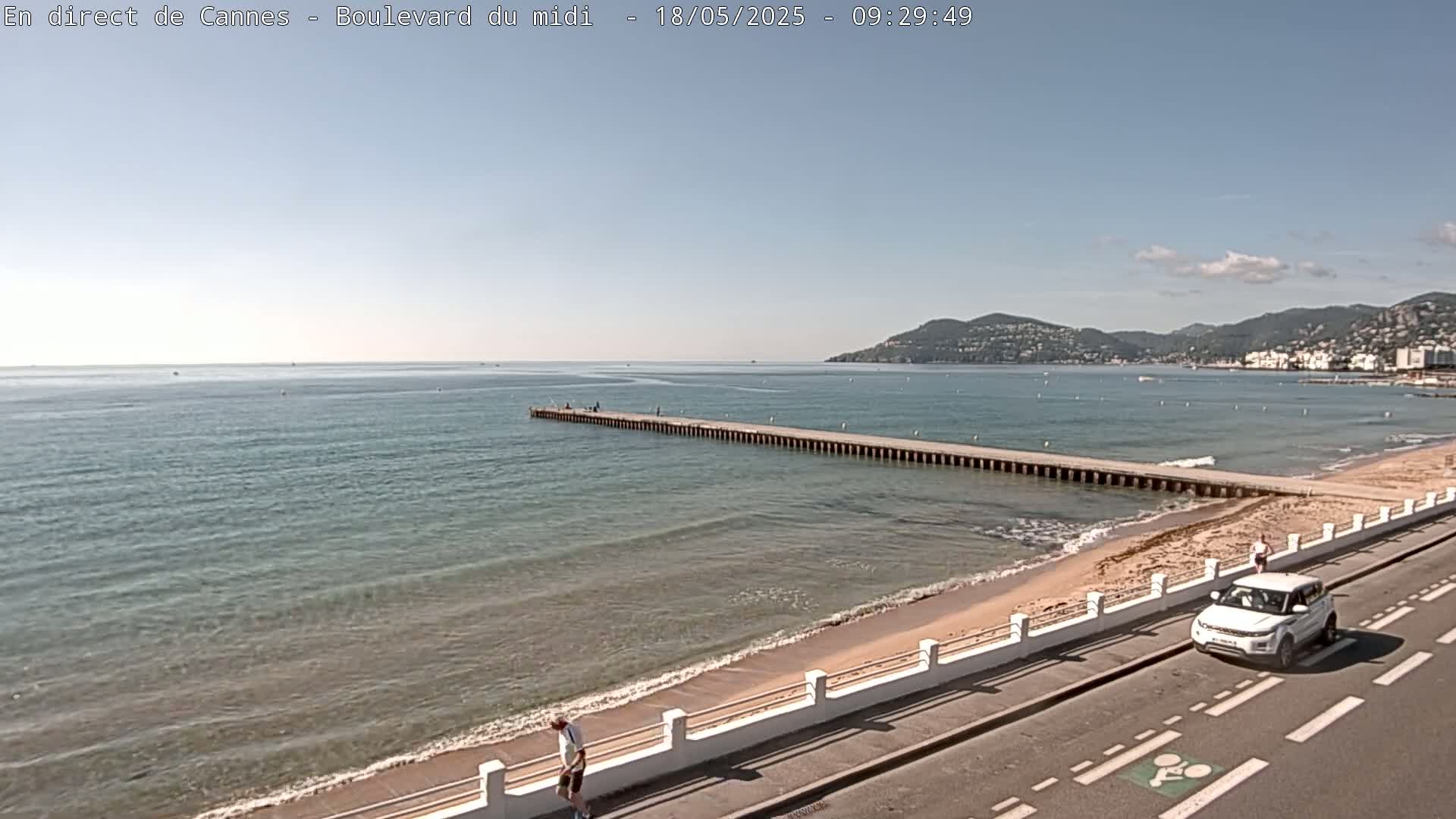 A long pier extends into a calm, blue-green sea under a clear, sunny sky, with a sandy beach and a road alongside it.
