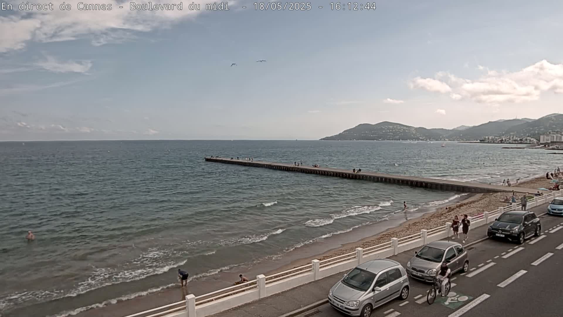 A partly cloudy day shows a long pier extending into a calm sea with a sandy beach and a road with cars parked along it, and people enjoying the beach.