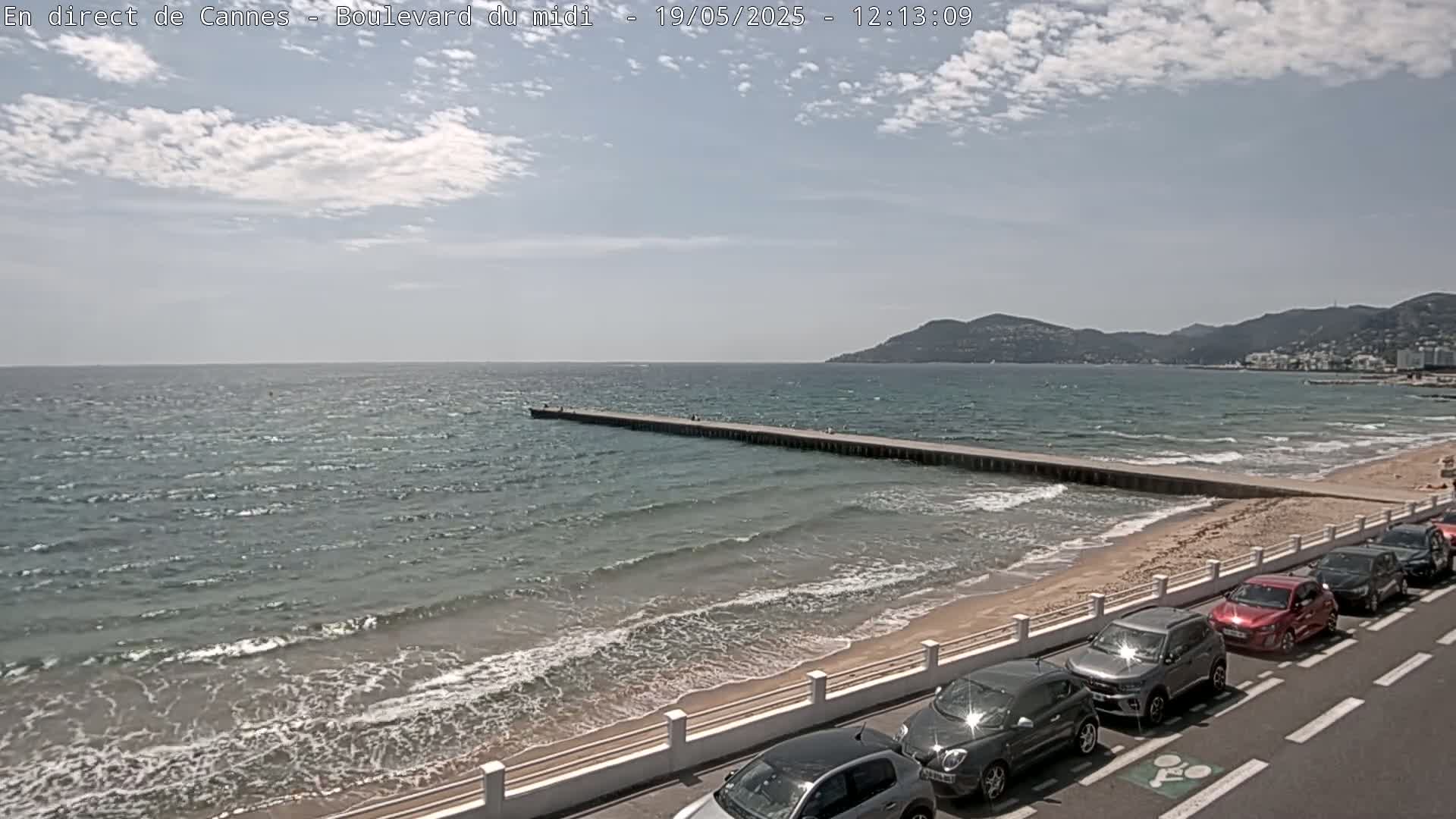 A sandy beach with a long pier extends into a moderately wavy sea under a partly cloudy sky, with several cars parked along the roadside.