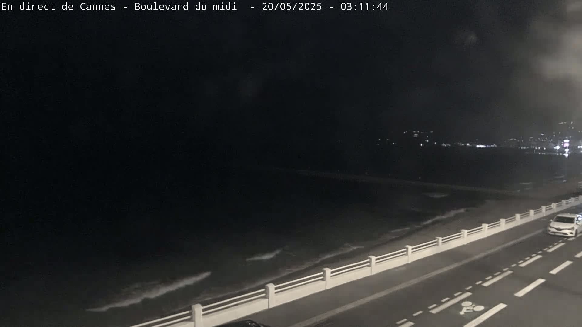 A dark nighttime scene shows a car driving along a coastal road with a low white barrier separating it from a dark ocean with small waves.
