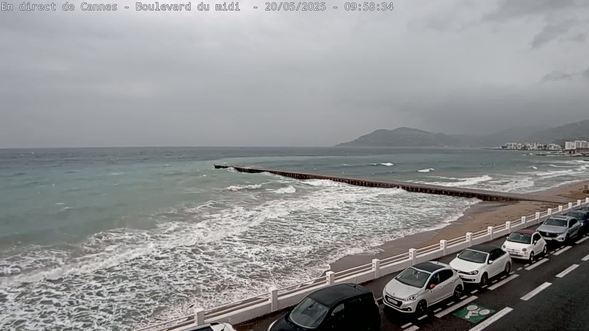 A beach with choppy waves under an overcast sky shows several cars parked along a roadside next to a low seawall.