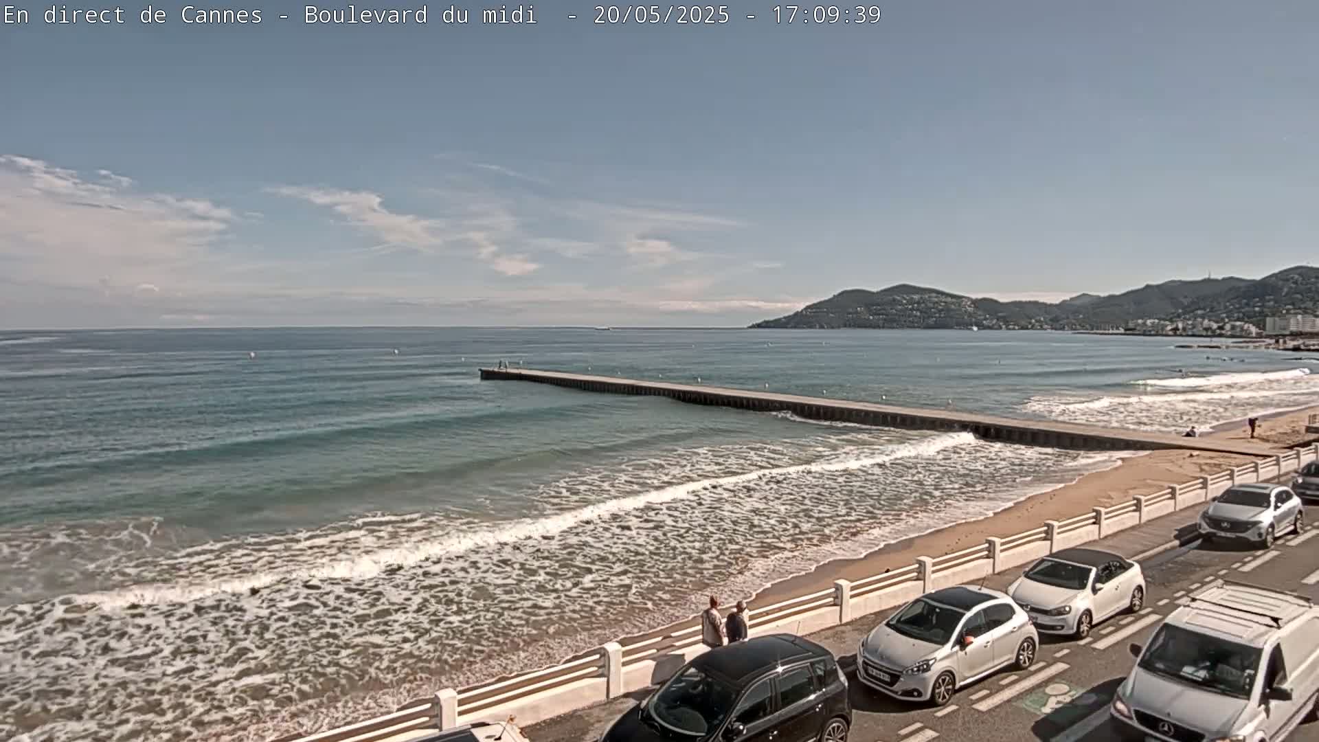 A sandy beach with gentle waves laps against a long pier extending into a calm sea under a partly cloudy sky, with several cars parked along the roadside.
