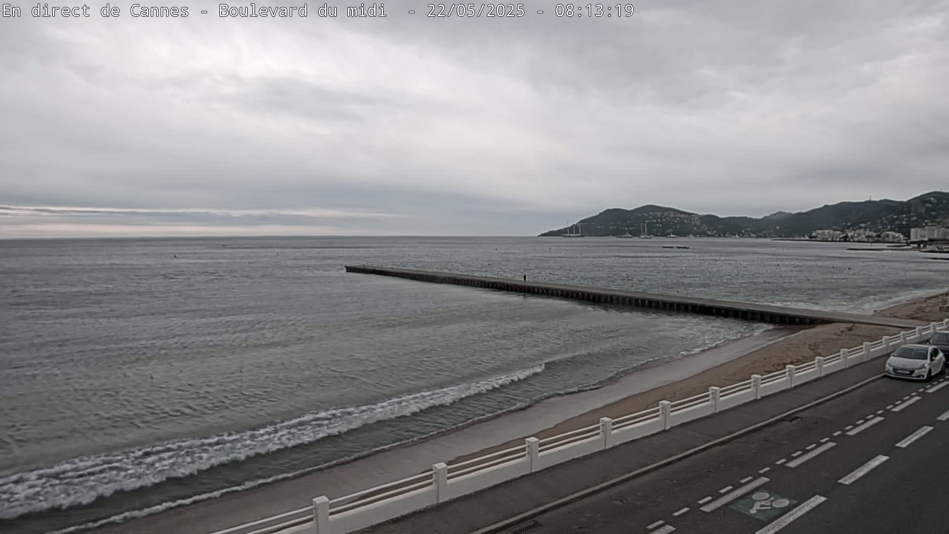 A long pier extends into a calm, gray sea with small waves lapping a sandy beach under an overcast sky, with hills visible in the distance and a road running alongside the shore.