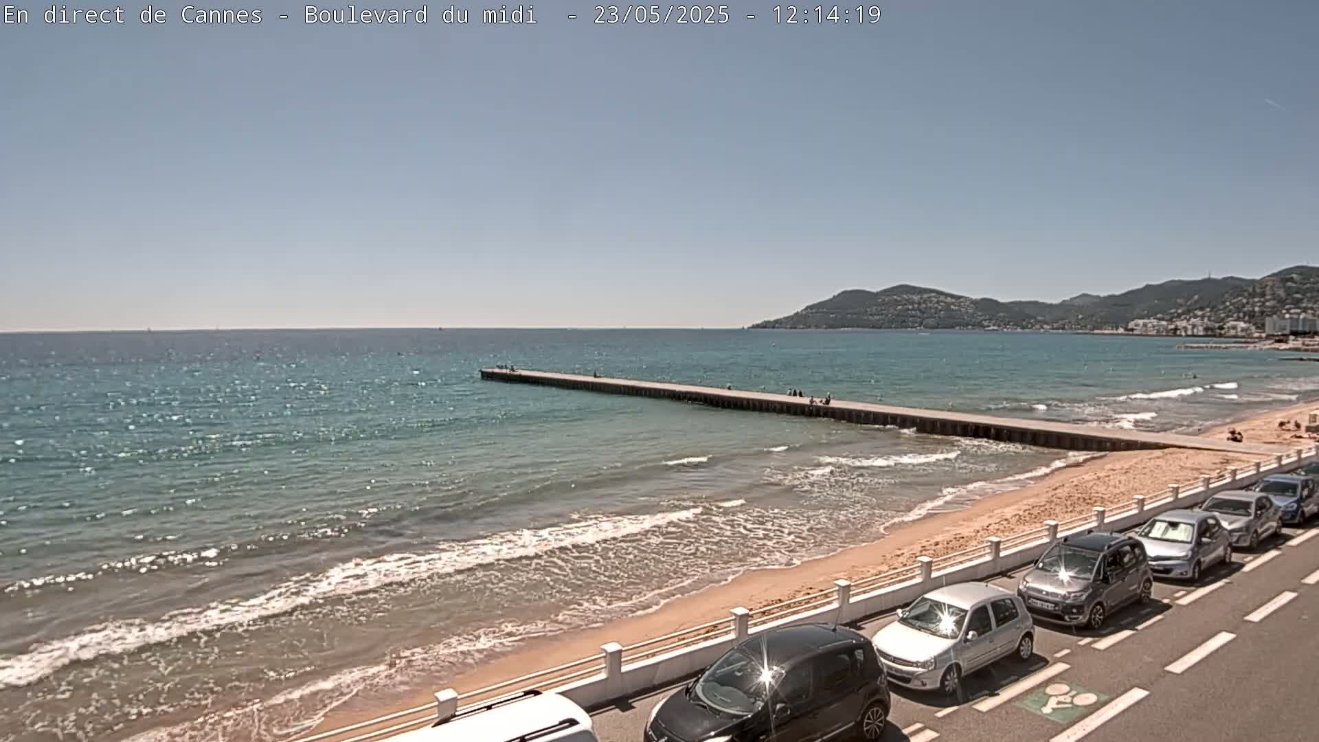 A long pier extends into a calm, blue ocean next to a sandy beach lined with parked cars under a clear sky.