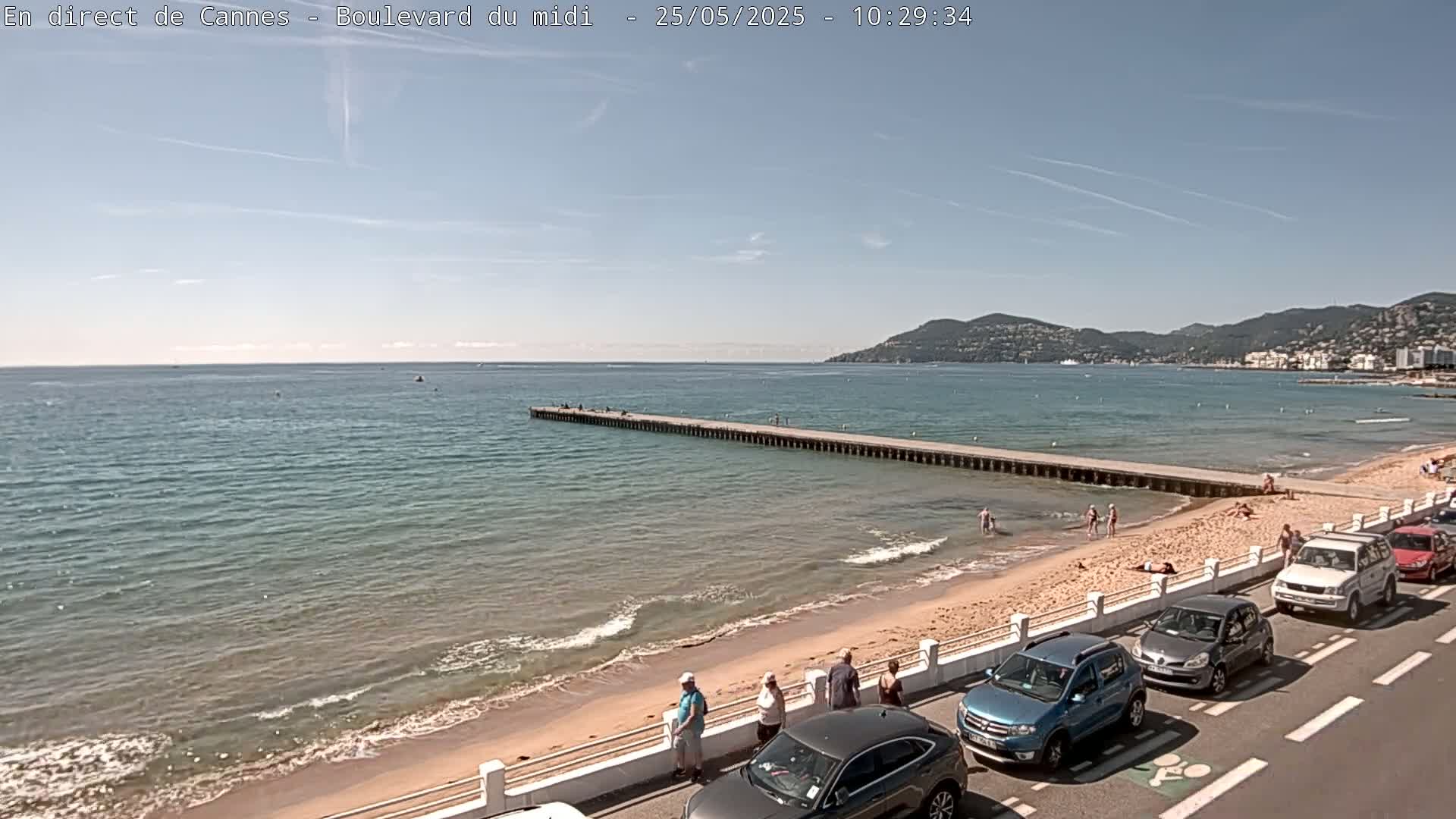 A sandy beach with a long pier extending into a calm, blue sea under a mostly clear sky, is lined with parked cars and a few people are visible on the beach and pier.