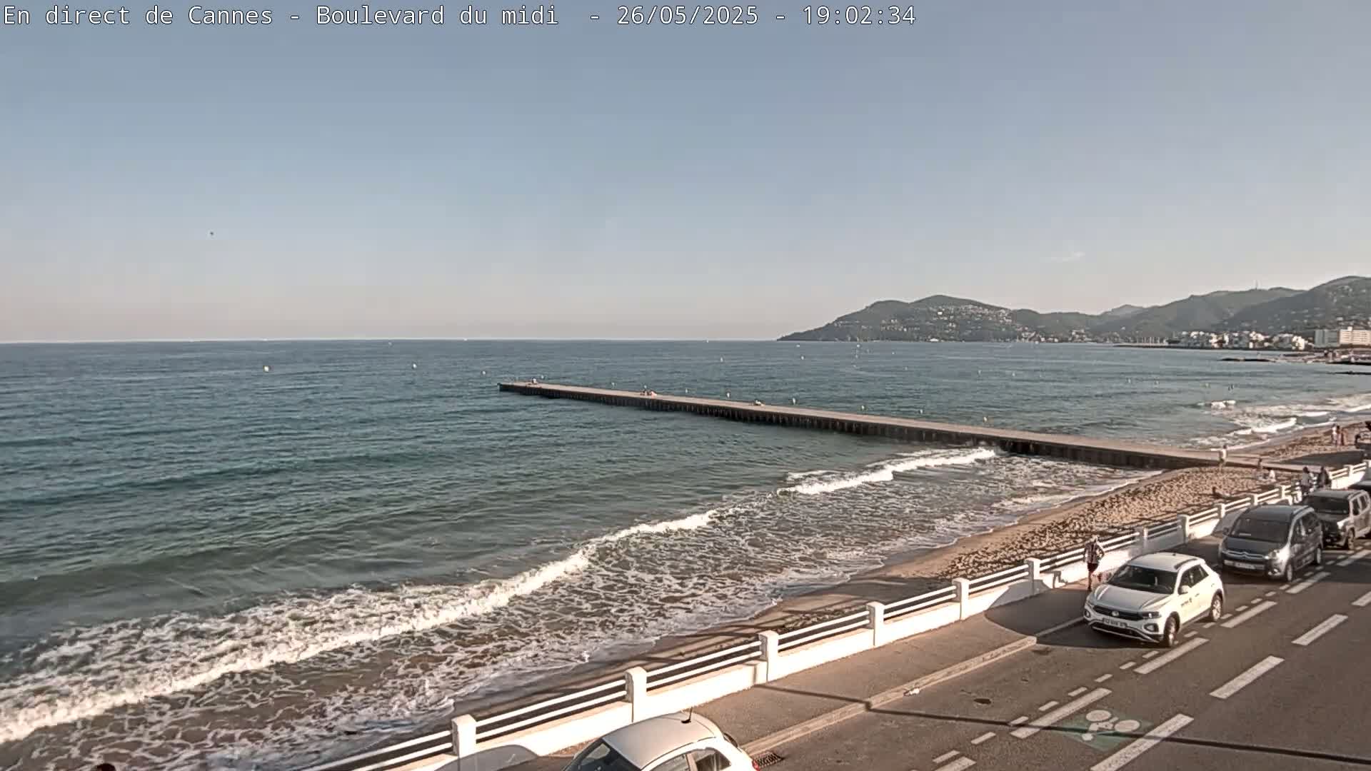 A long pier extends into a calm sea with small waves lapping a sandy beach, backed by a road with parked cars and distant hills under a clear sky.