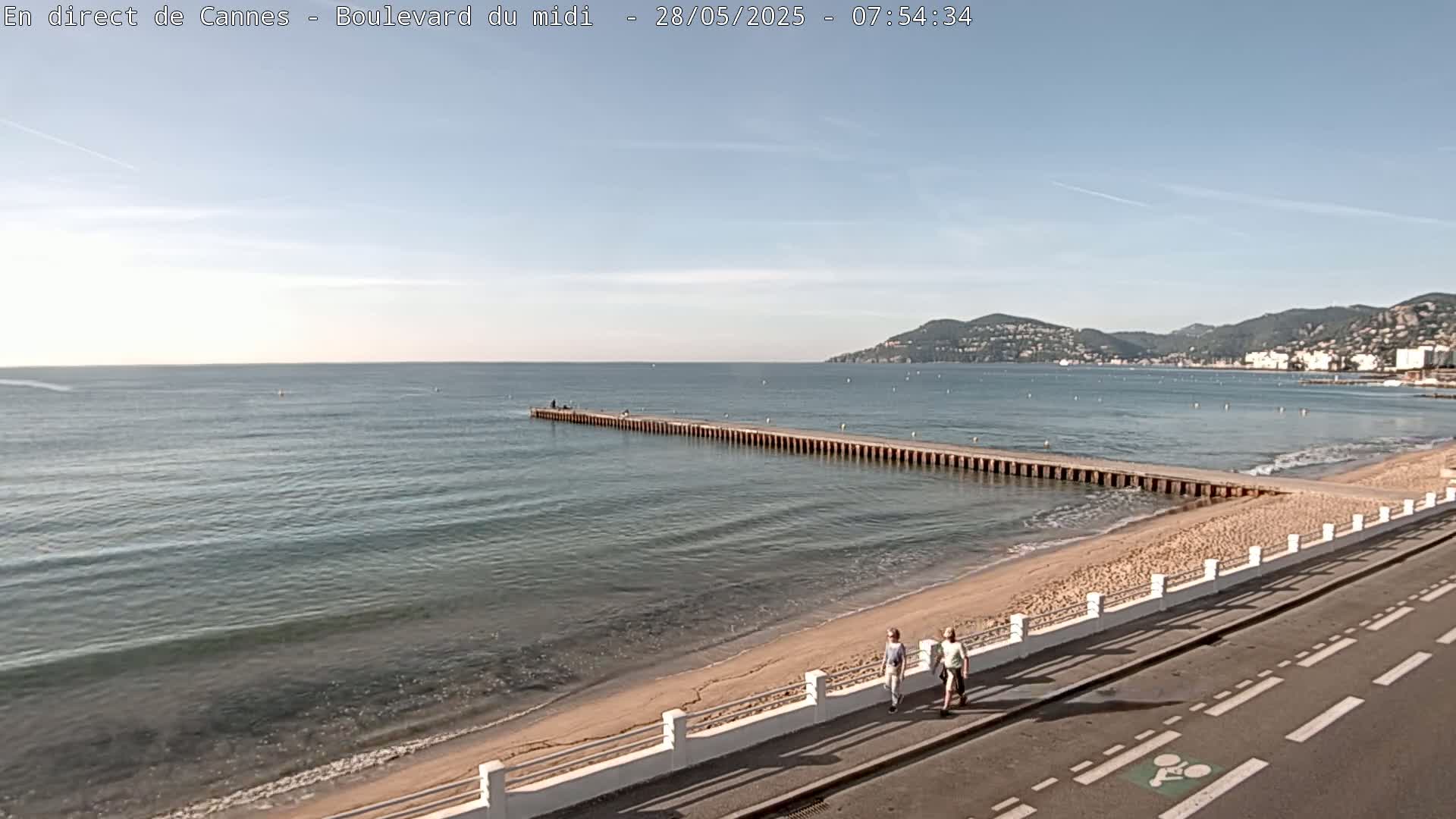 A long pier extends into a calm sea on a sunny day, with a sandy beach and two people walking along a road next to it, all backed by distant hills.
