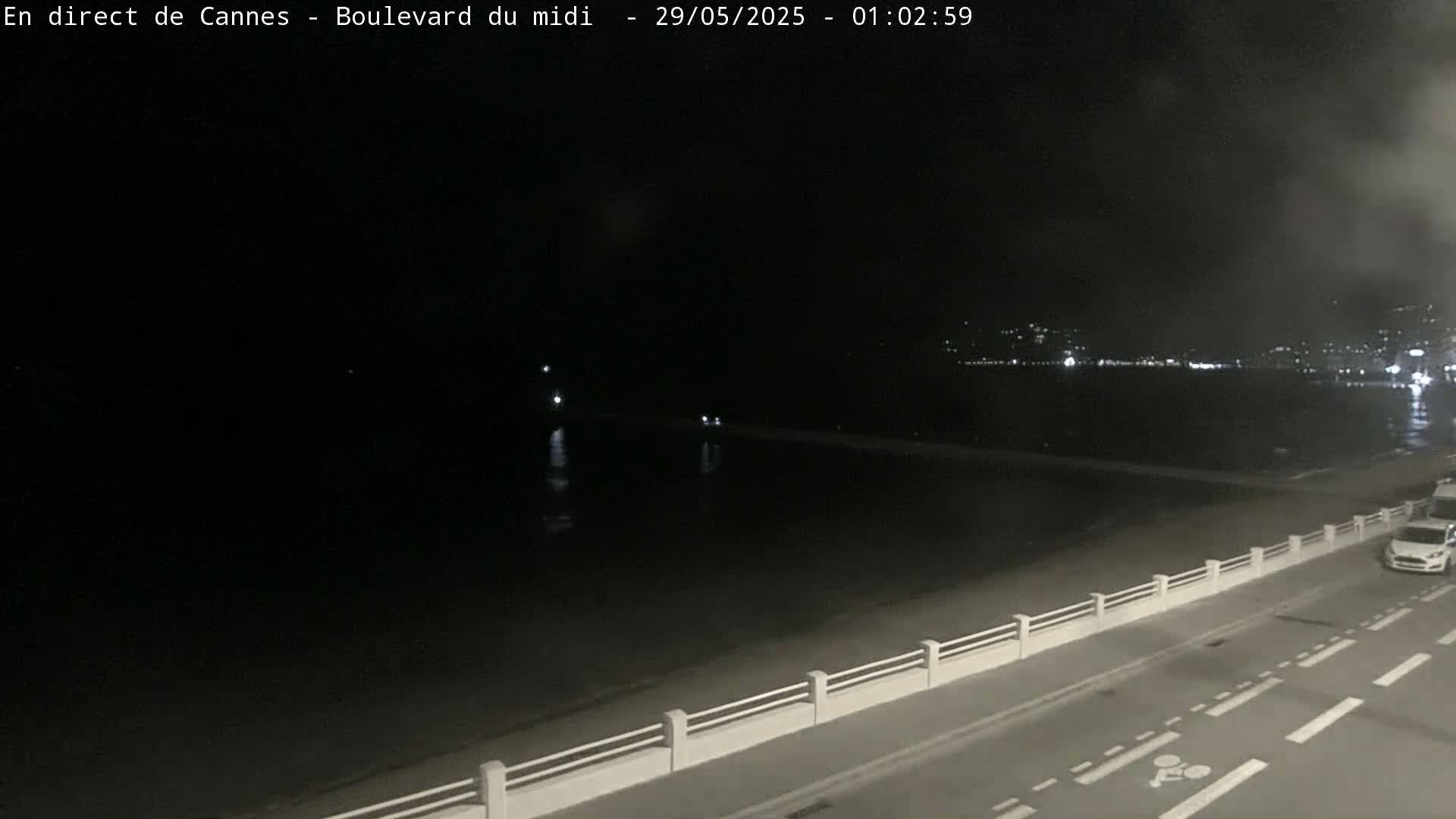 A nighttime view of a calm body of water next to a road with a low white fence, and a few cars visible on the road.