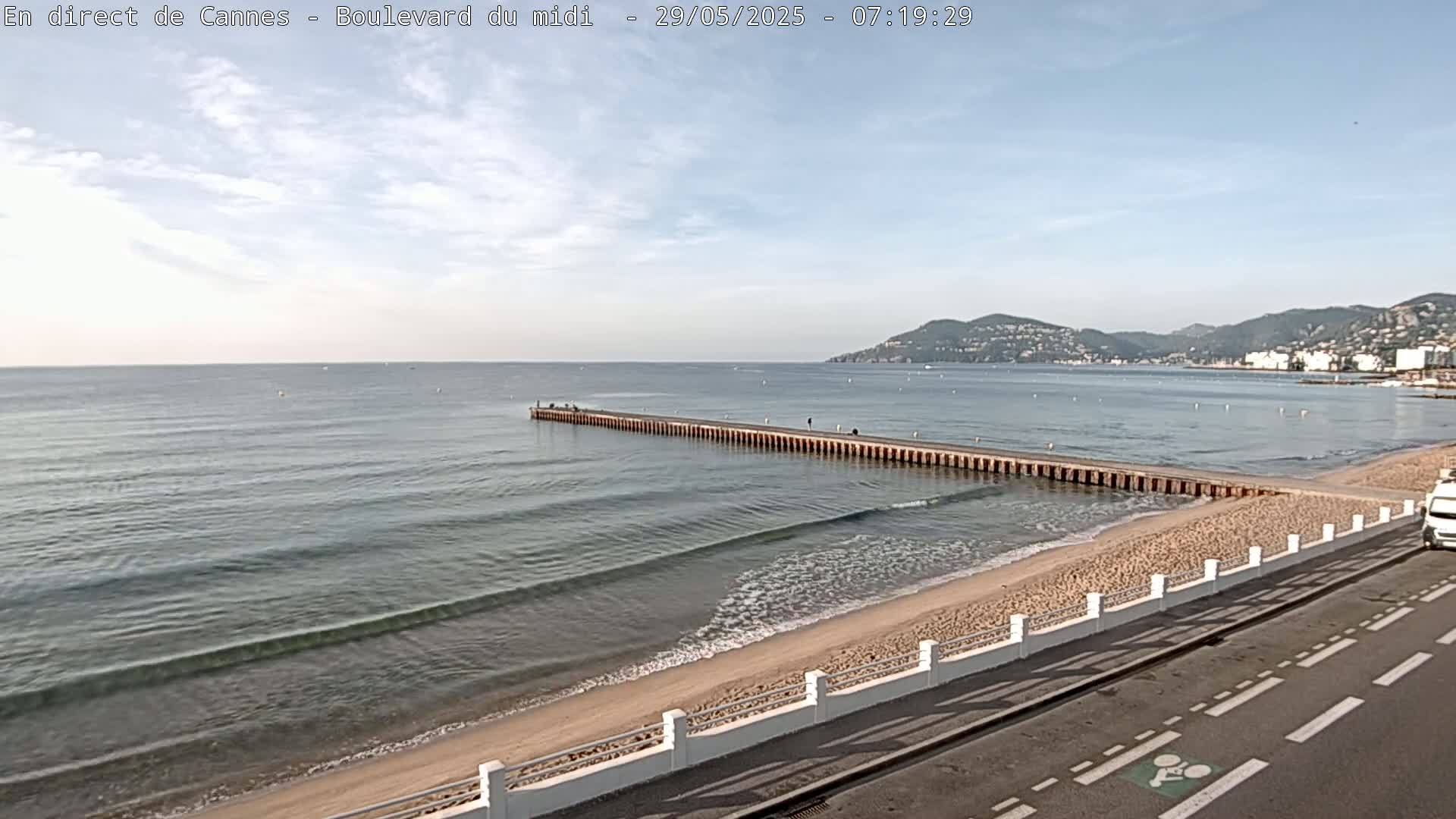 A long pier extends into a calm sea with small waves lapping a sandy beach under a mostly clear sky, backed by distant hills.