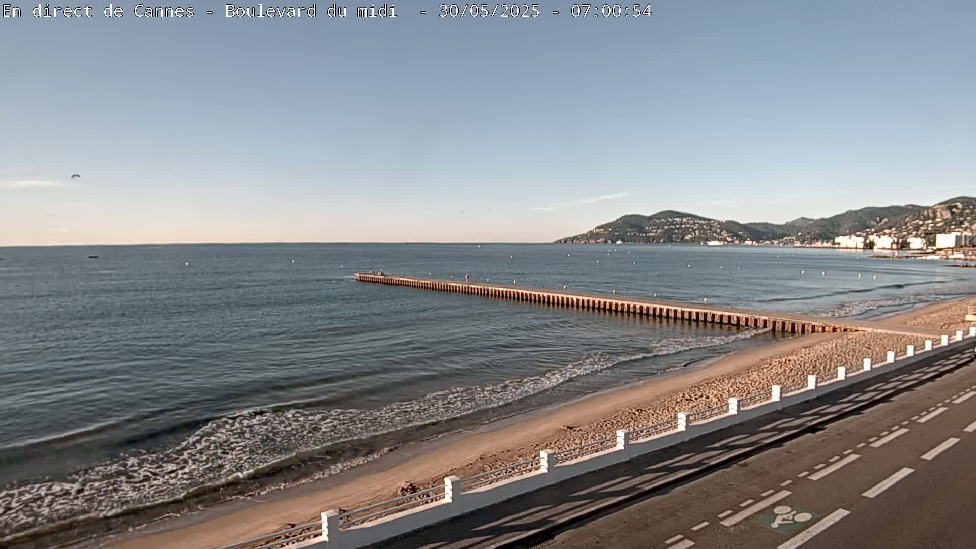 A long pier extends into a calm, blue sea, alongside a sandy beach and a paved road under a clear sky.