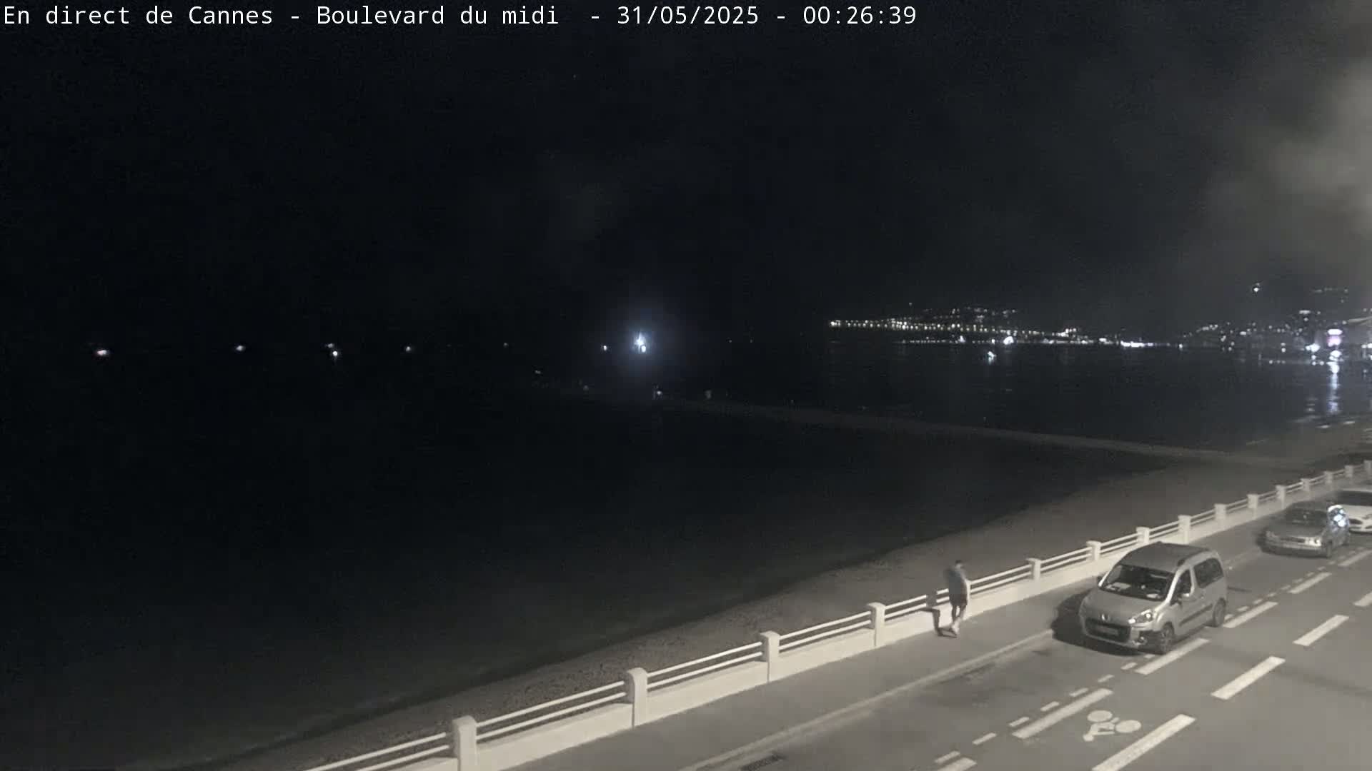 A nighttime view of a mostly dark beach and roadway next to a body of water, with a person walking along the road and several cars parked nearby, under a clear night sky.
